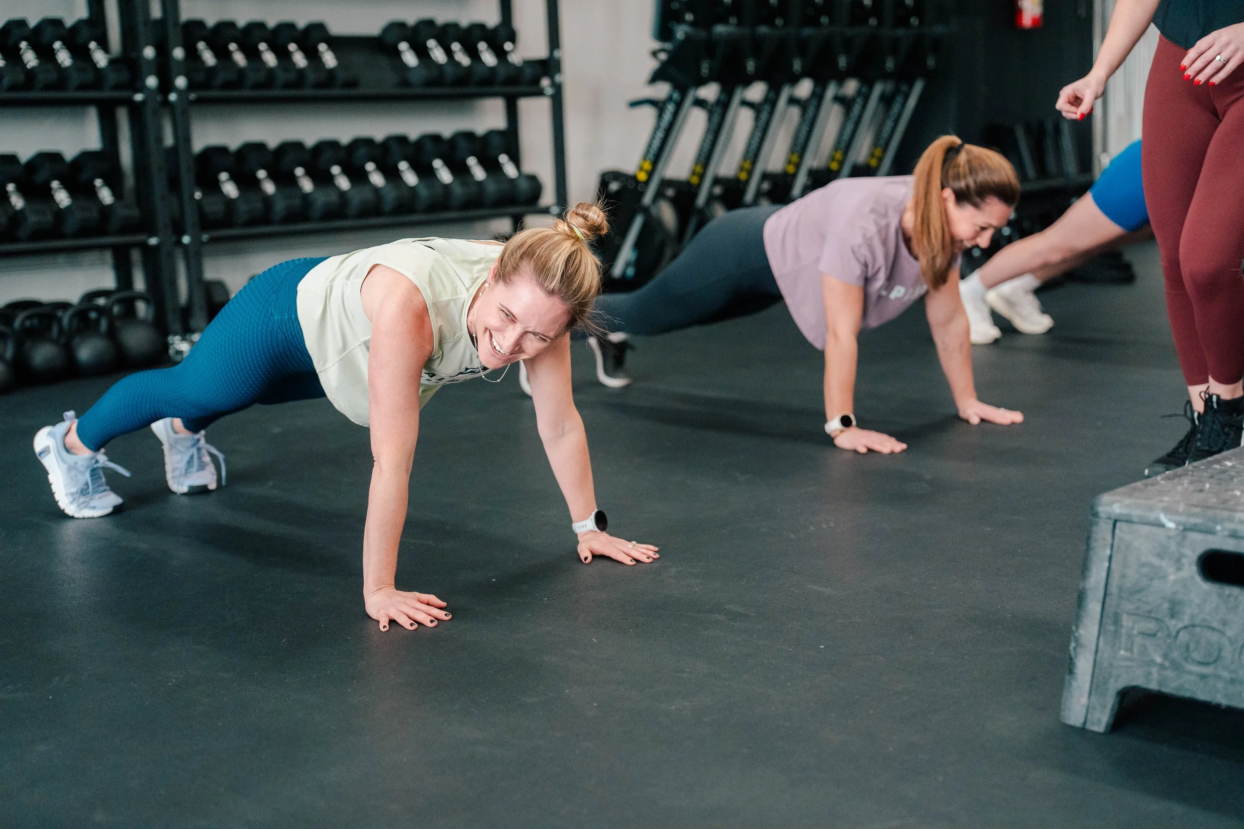 Women doing push-ups in a gym, smiling, with workout equipment in the background.