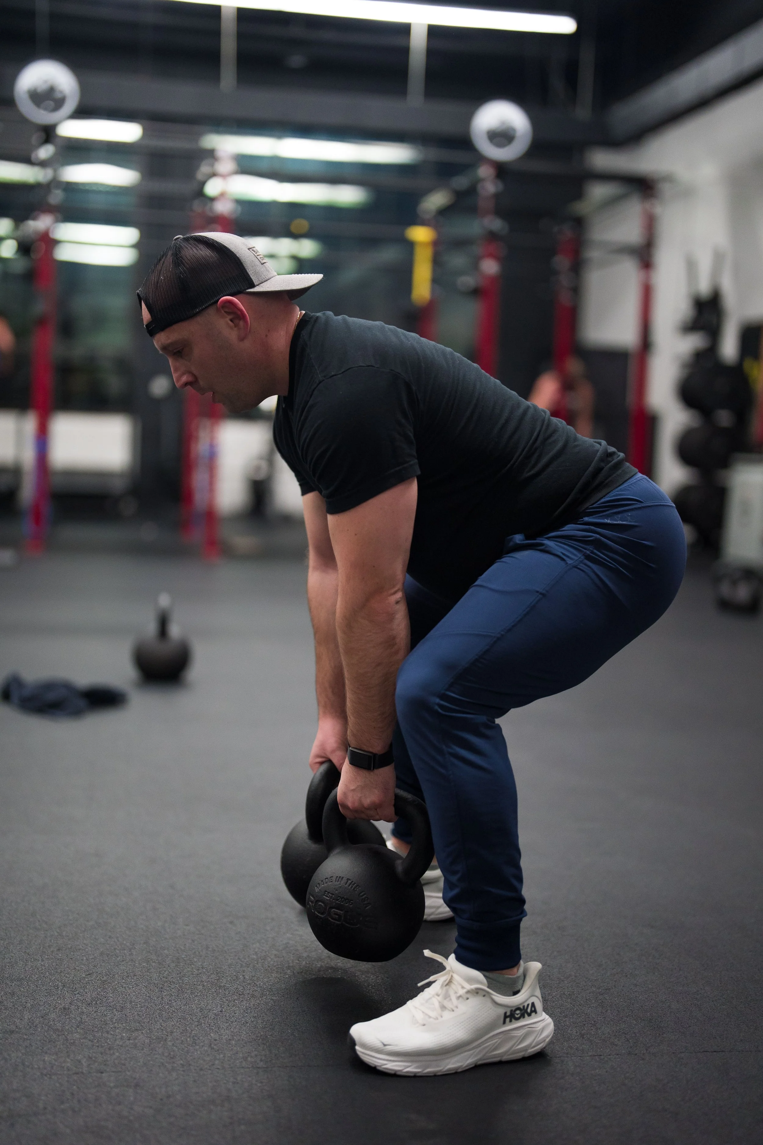 A man wearing a black t-shirt, blue pants, and white sneakers is performing a deadlift with a kettlebell in a gym.