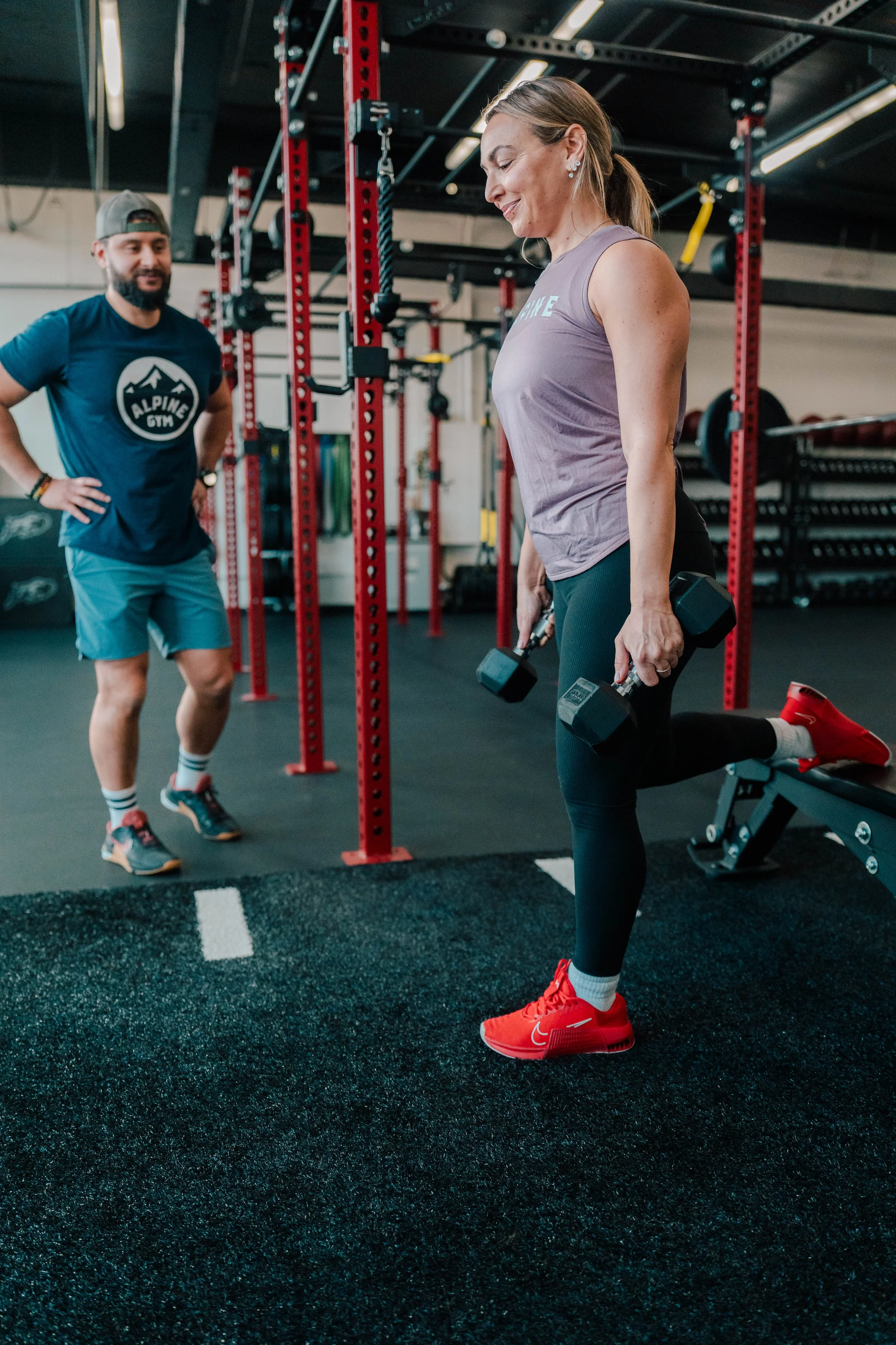 A woman in workout clothes lifting dumbbells during a workout session at a gym, with a male trainer watching her in the background.