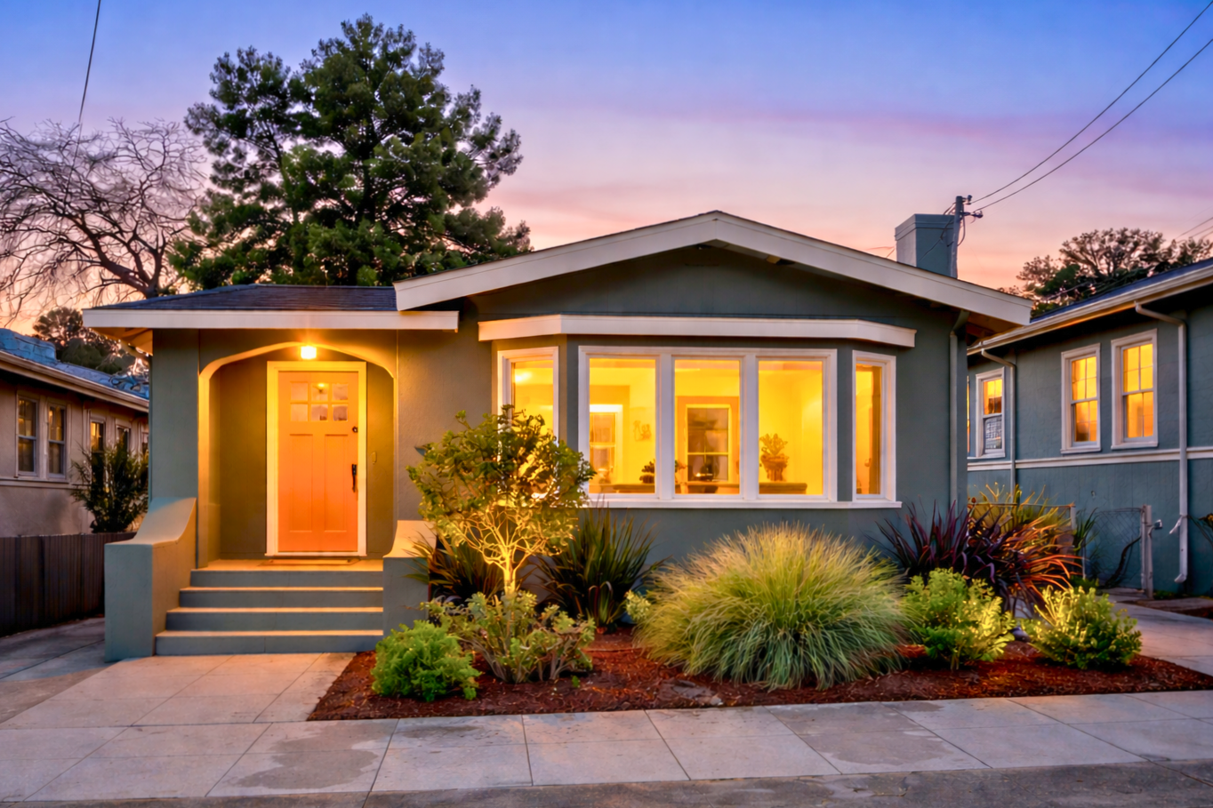 A cozy house at dusk with warm lit windows revealing a welcoming interior, green plants and shrubs in the front yard, concrete steps leading to a front door, with a large tree and power lines in the background.