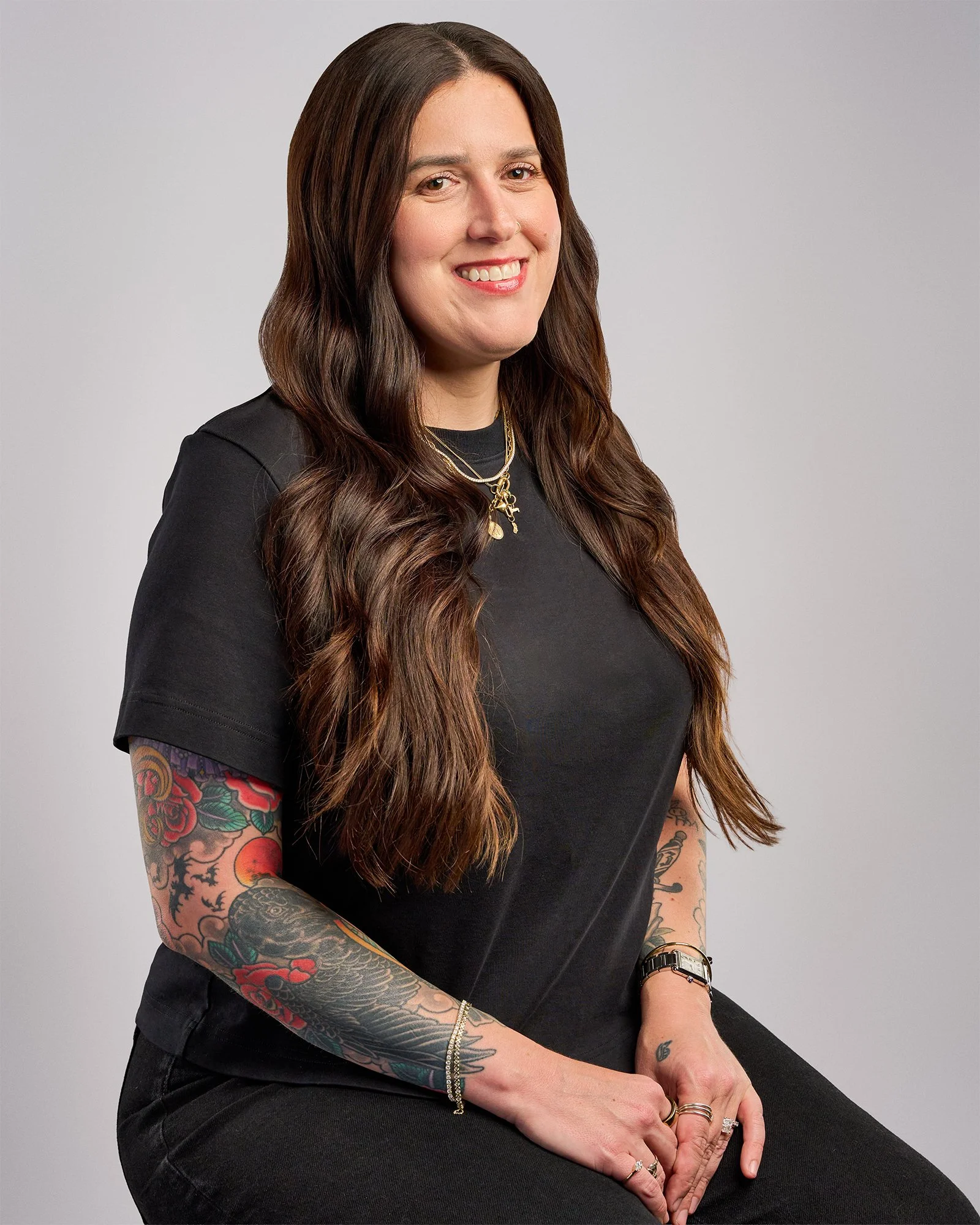A woman with long, wavy brown hair, tattoos on her arms, wearing a black shirt and jewelry, sitting against a plain gray background.