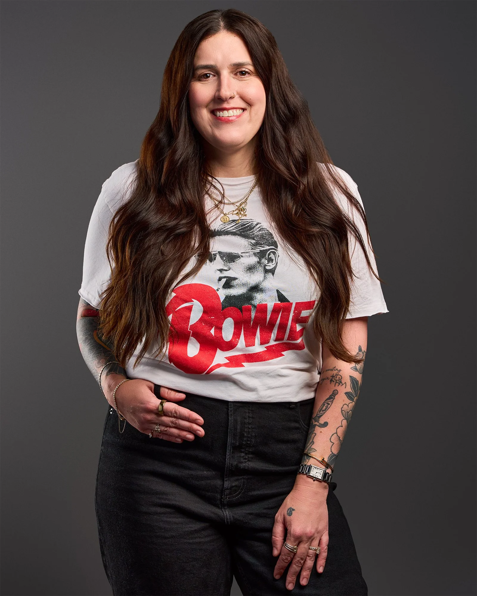 A woman with long dark hair, wearing a white graphic T-shirt, black jeans, multiple rings, a wristwatch, and layered gold necklaces, standing against a dark gray background.