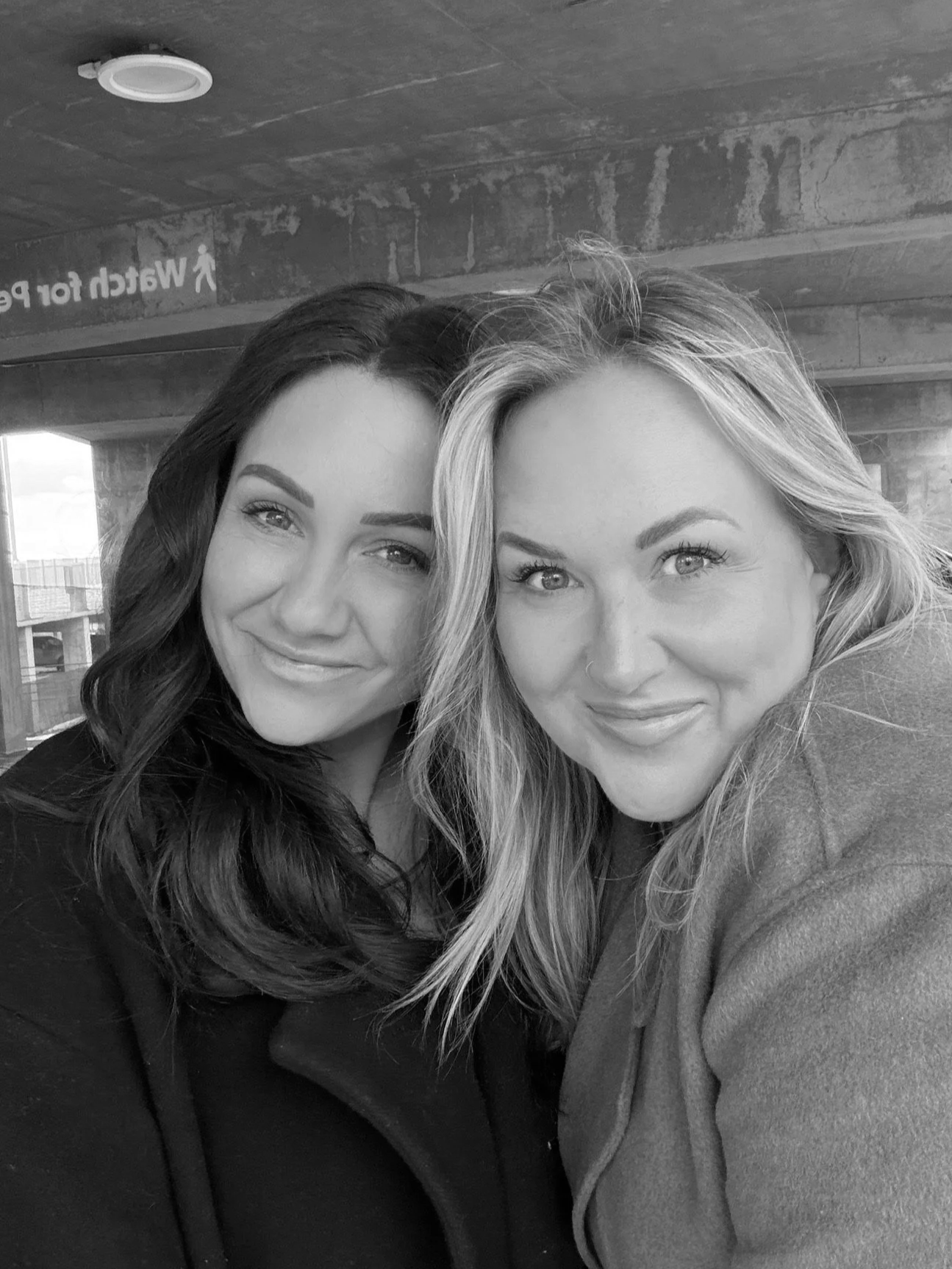 Two women taking a selfie together, smiling, inside a building with concrete walls. One woman has long, wavy dark hair and the other has long, wavy blonde hair.