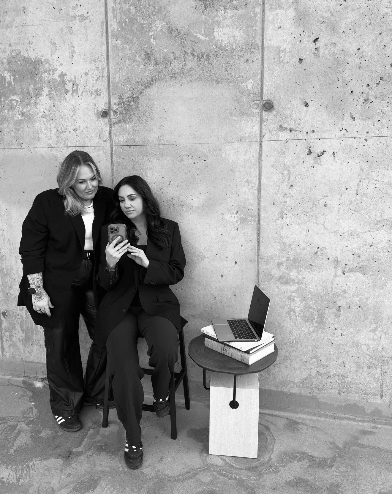 Two women in dark suits look at a phone together, with a laptop and books on a small table beside them, set against a concrete wall.