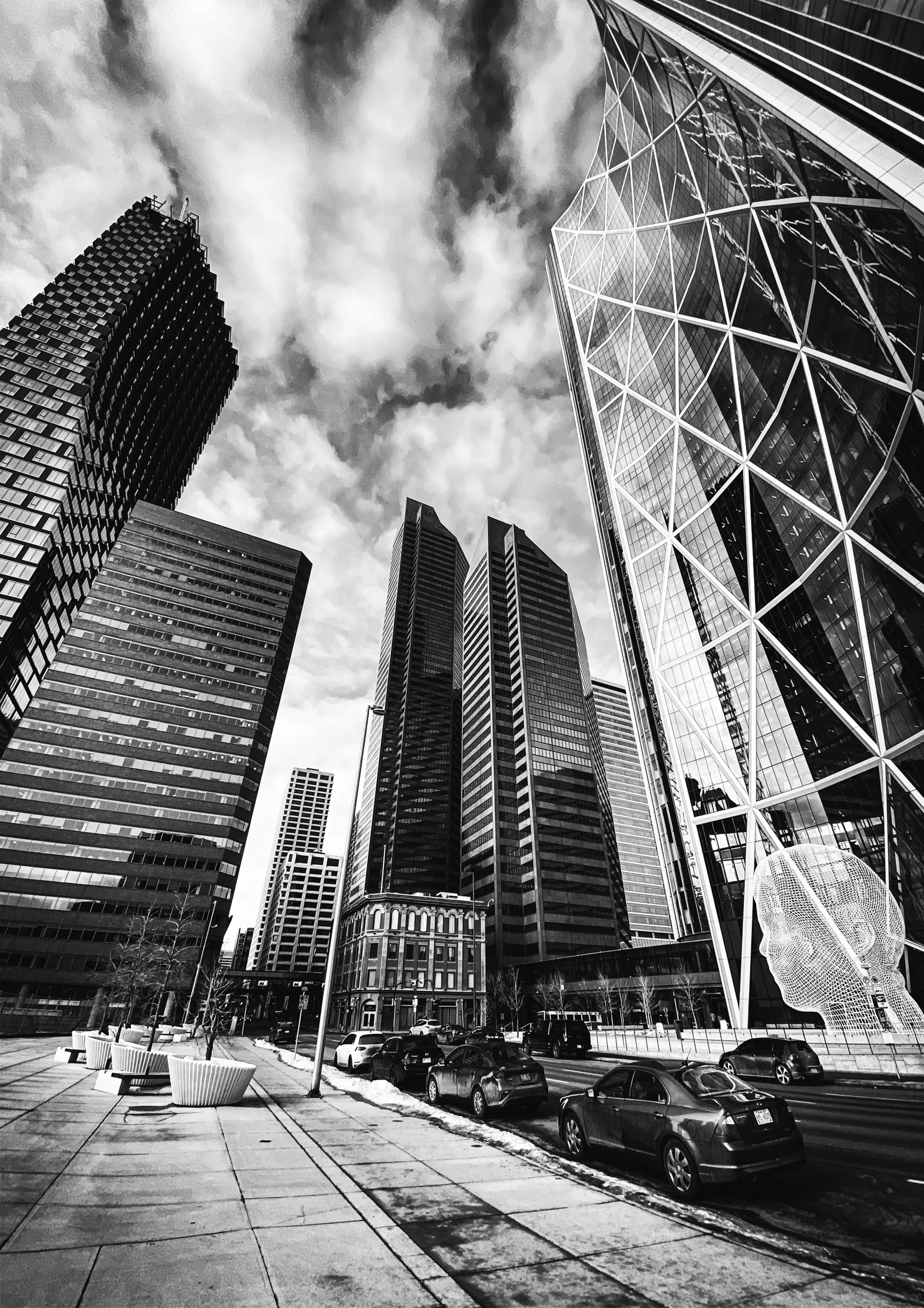 Black and white photo of tall modern skyscrapers with glass facades, parked cars on street, and clouds in sky.