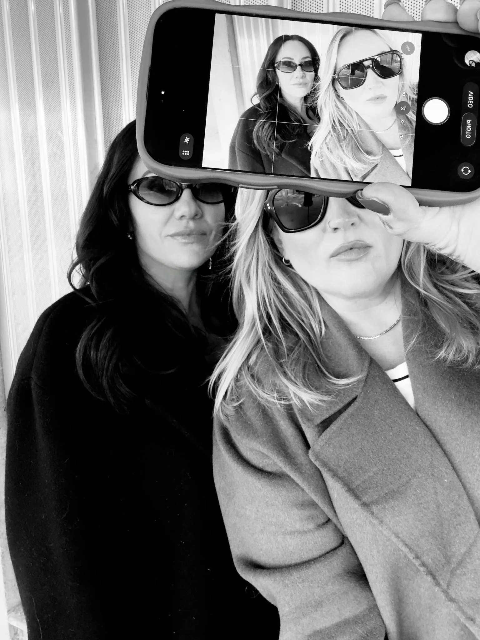 Two women taking a selfie together, wearing sunglasses, with one woman holding a smartphone in front of the other. The photo is being captured on the phone's screen visible in the foreground.