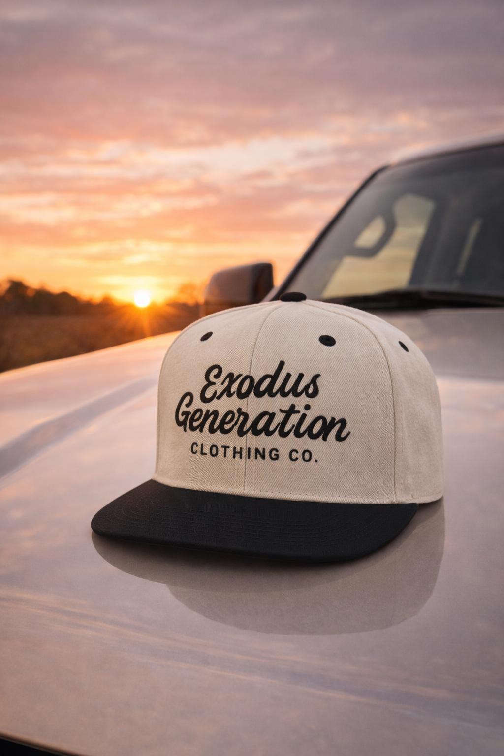 A baseball cap with the text "Exodus Generation Clothing Co." on the front, resting on the hood of a vehicle during a sunset with a colorful sky.