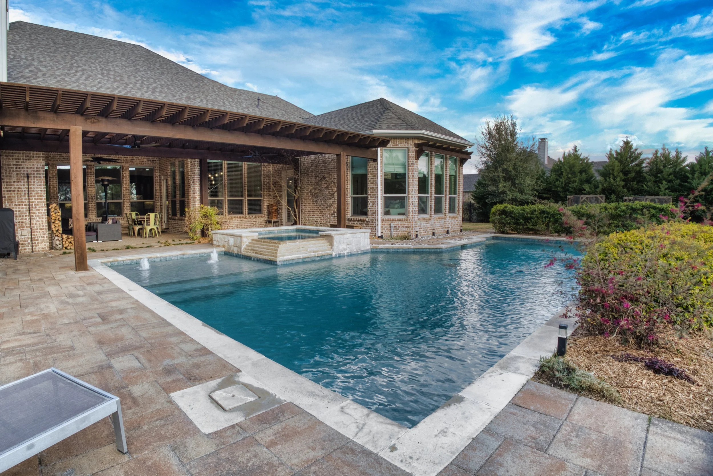 Backyard with a swimming pool, hot tub, brick house, and outdoor seating area under a covered patio, surrounded by trees and shrubs under a partly cloudy sky.