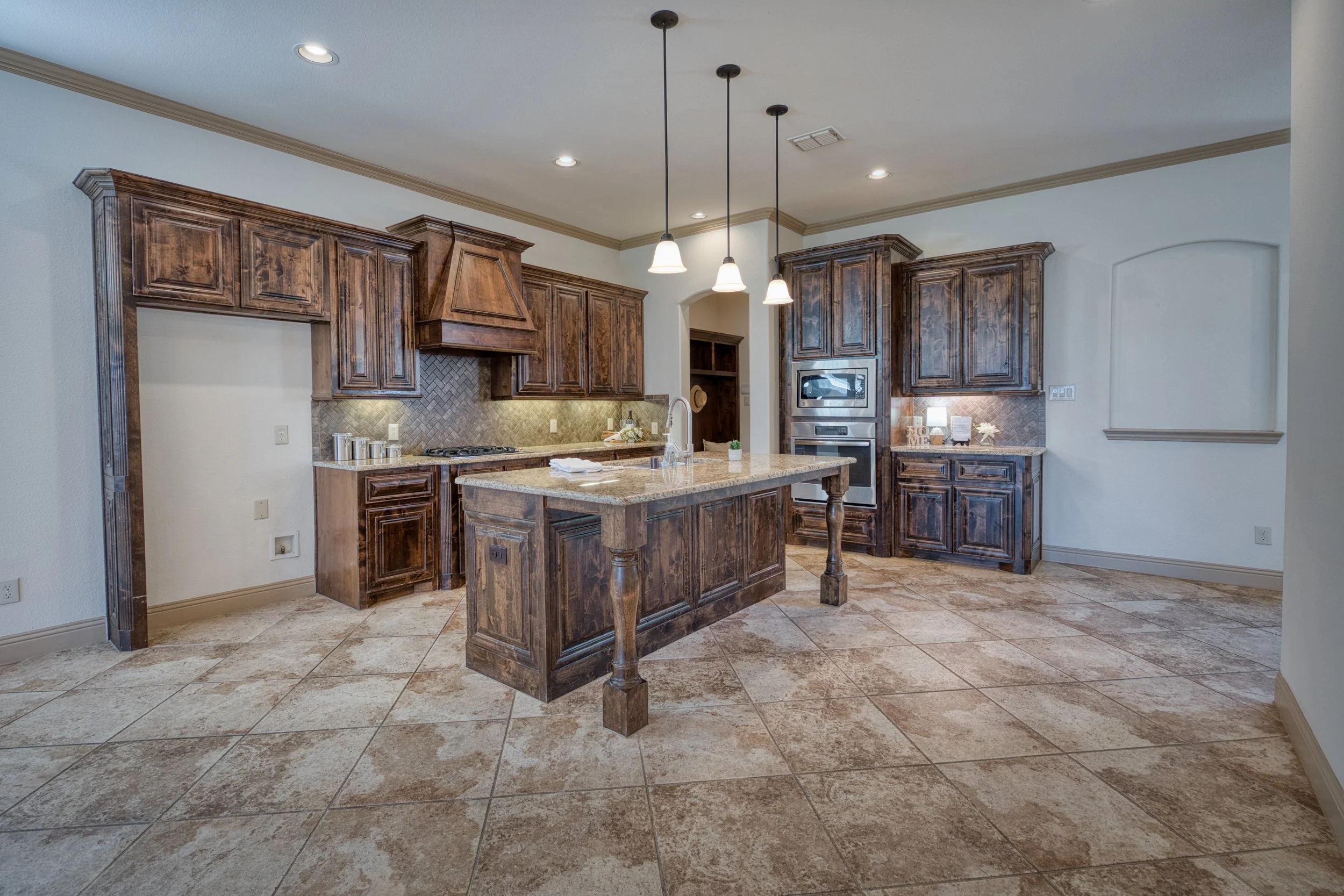 Kitchen with dark wood cabinets, a kitchen island, and beige tiled floors. Pendant lights hang above the island, and there are built-in appliances and a backsplash with a herringbone pattern.