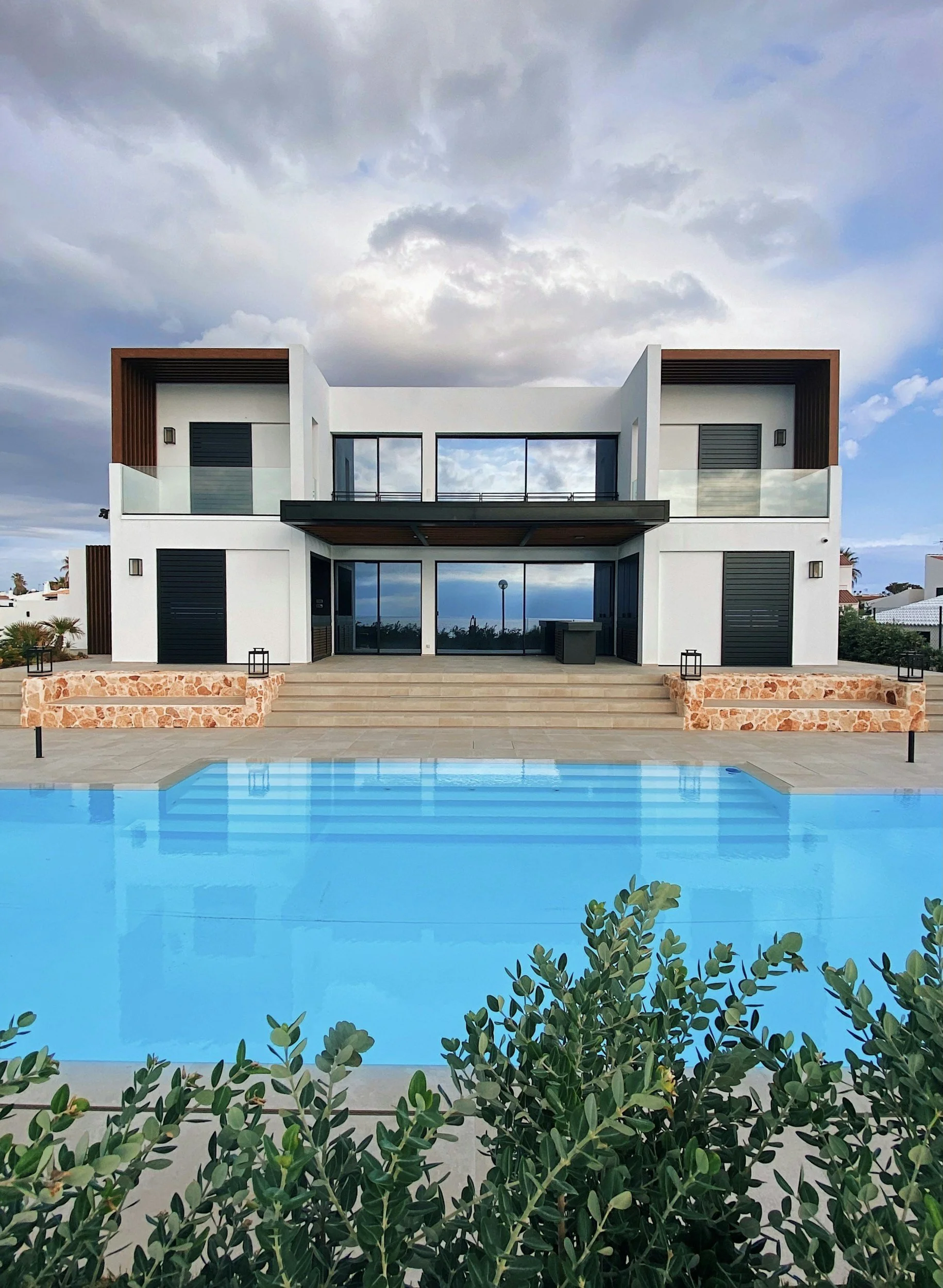 Modern two-story house with large glass doors, a swimming pool in the foreground, and overcast sky.