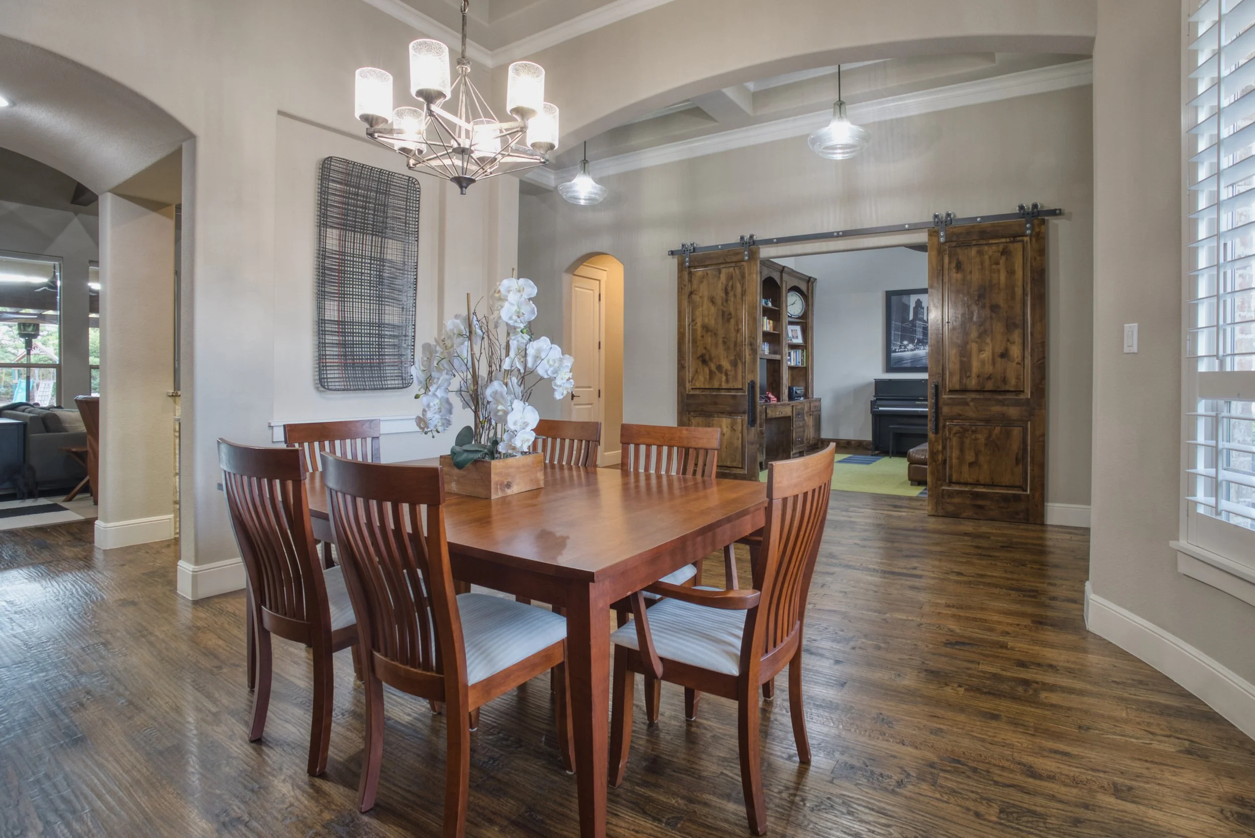 A dining room with a wooden table and six chairs, a rectangular wall art piece, a chandelier, and a sliding barn door leading to a living room with a piano and framed artwork.