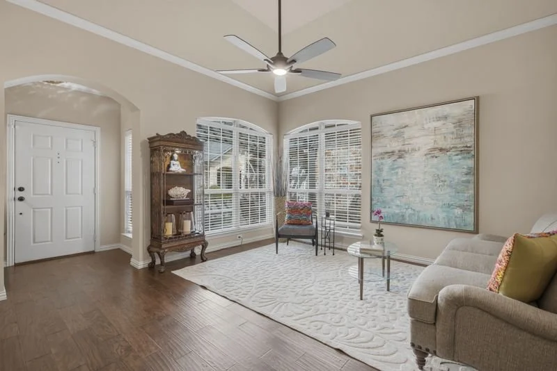 Living room with large arched windows, a ceiling fan, a beige couch with pillows, a glass coffee table, a colorful armchair, an ornate wooden display cabinet, a large abstract wall art, and a white textured rug.