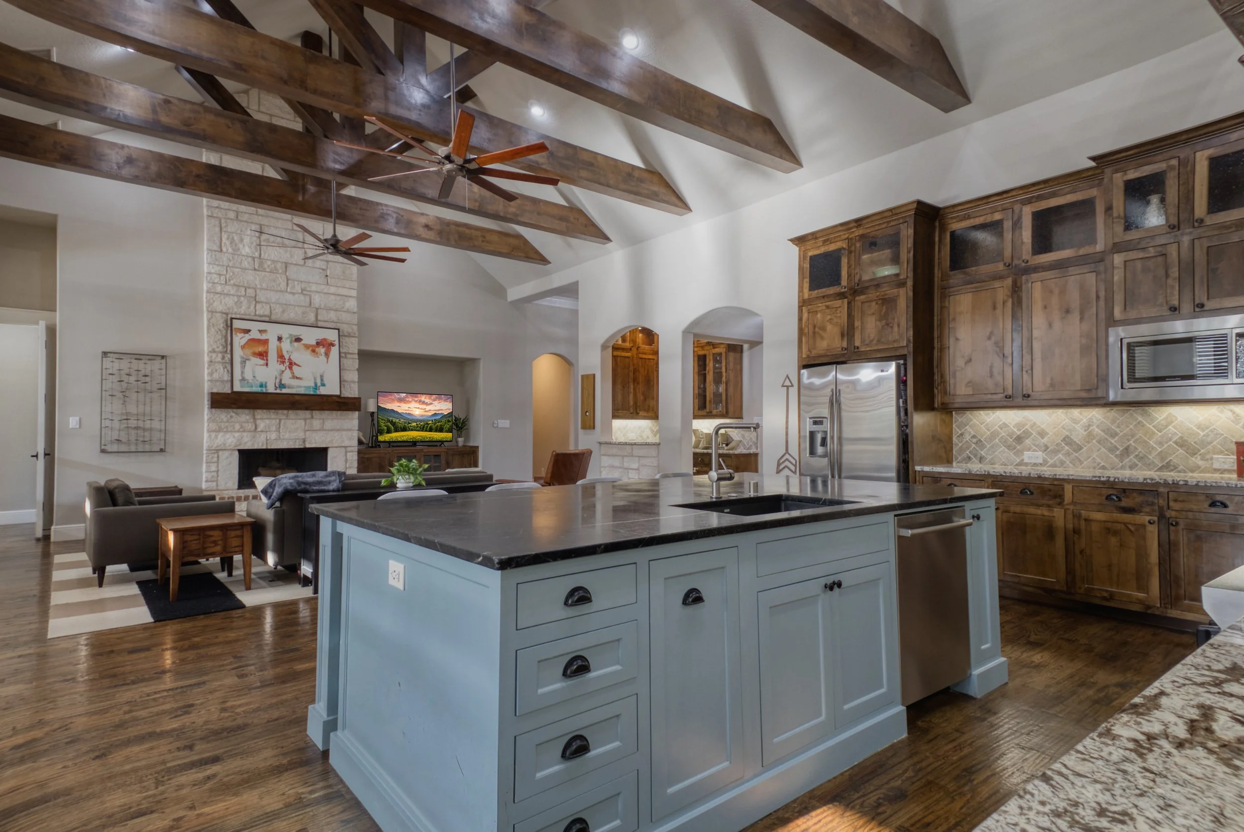 Open-concept kitchen and living room with vaulted ceilings, exposed wooden beams, hardwood floors, a central kitchen island with a black countertop, wooden cabinets, and a stone fireplace in the living area with a TV above it.