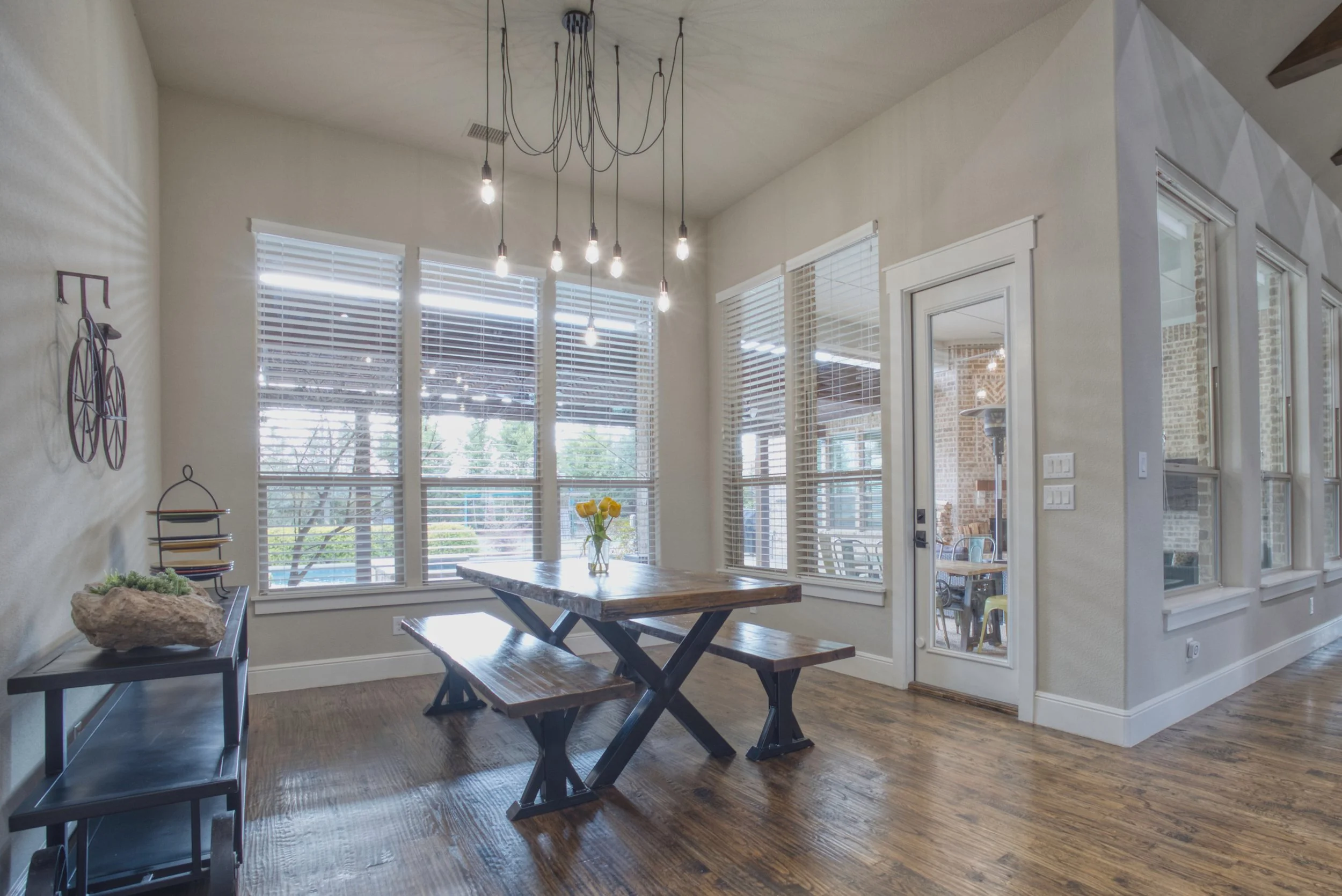 Dining area with wooden table and benches, multiple windows with white blinds, a decorative chandelier with exposed bulbs, and a door leading outside, with hardwood flooring and minimal decor.