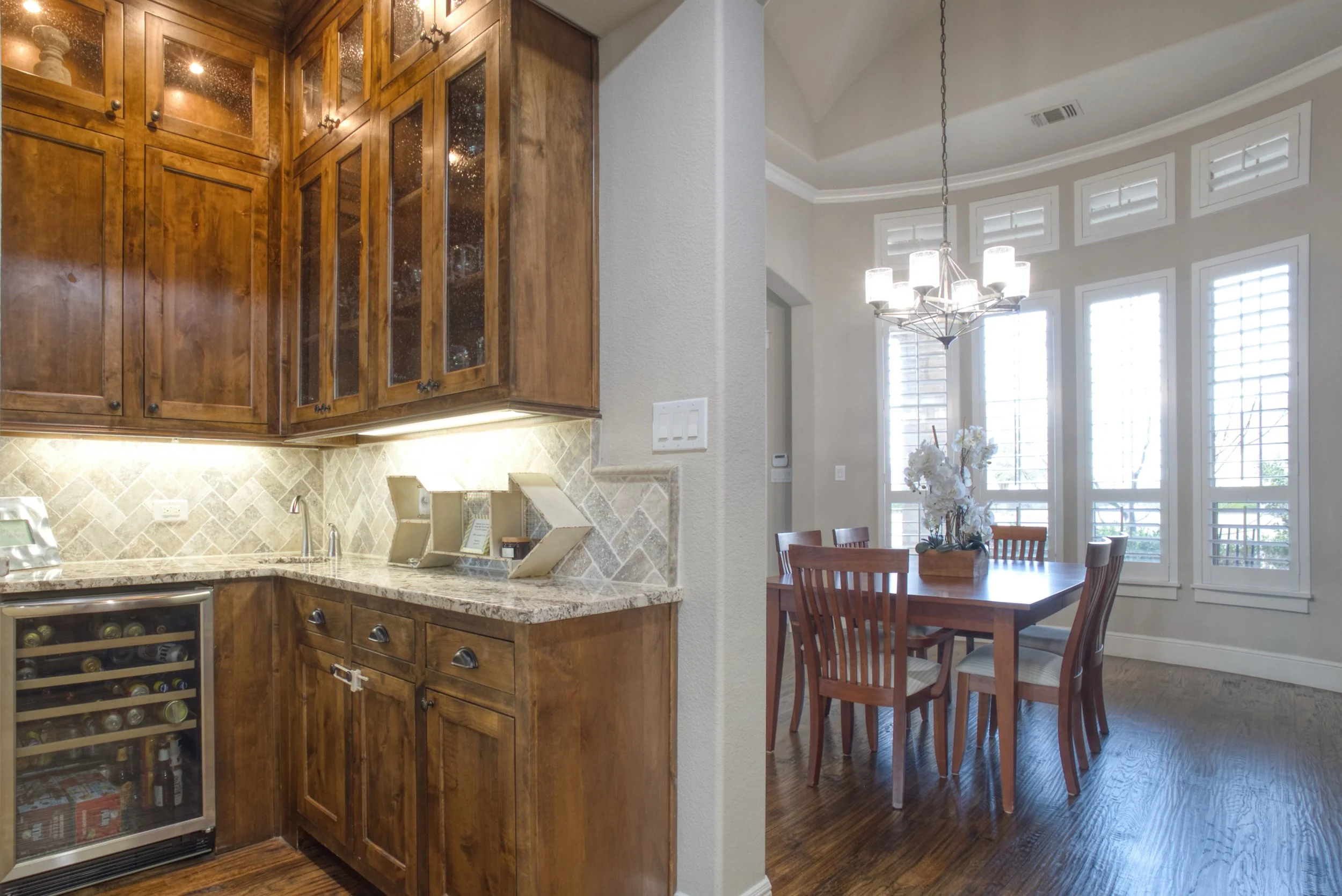 Part of a kitchen with wooden cabinets, a granite countertop, and a wine cooler, adjacent to a dining area with a wooden table, chairs, and large windows with white shutters, and a chandelier hanging from the ceiling.
