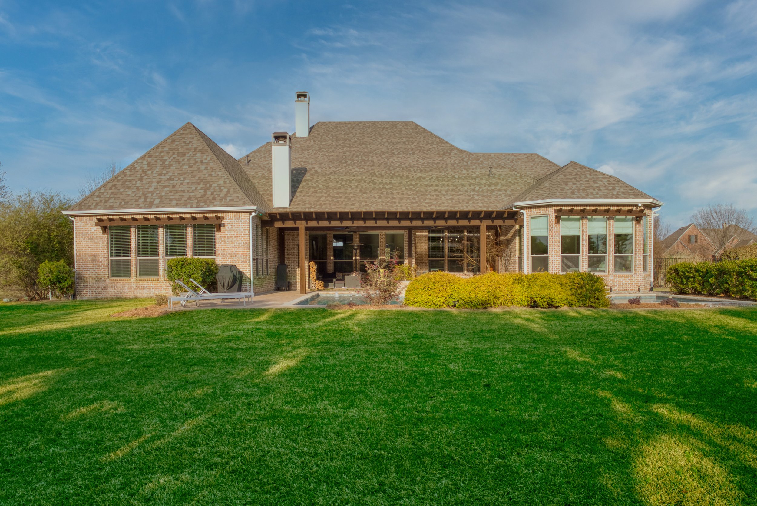 Rear view of a large brick house with a brown shingle roof, multiple chimneys, large windows, a covered patio area, a swimming pool, lounge chairs, green lawn, bushes, and other neighboring houses in the background.