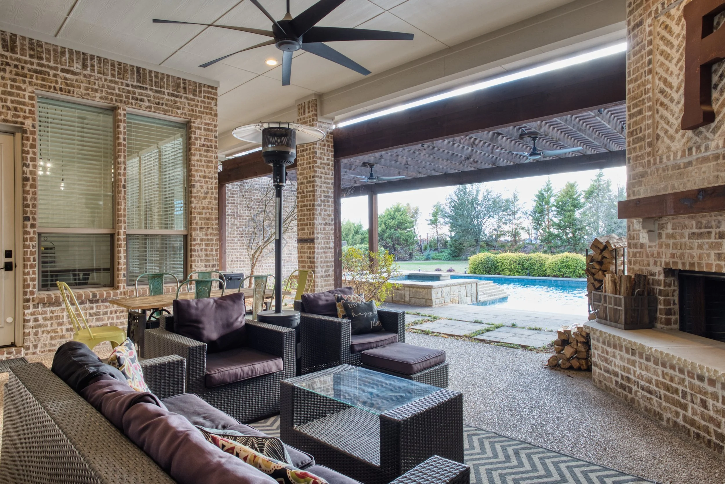 Covered outdoor patio with seating area, fireplace, and view of pool and garden.