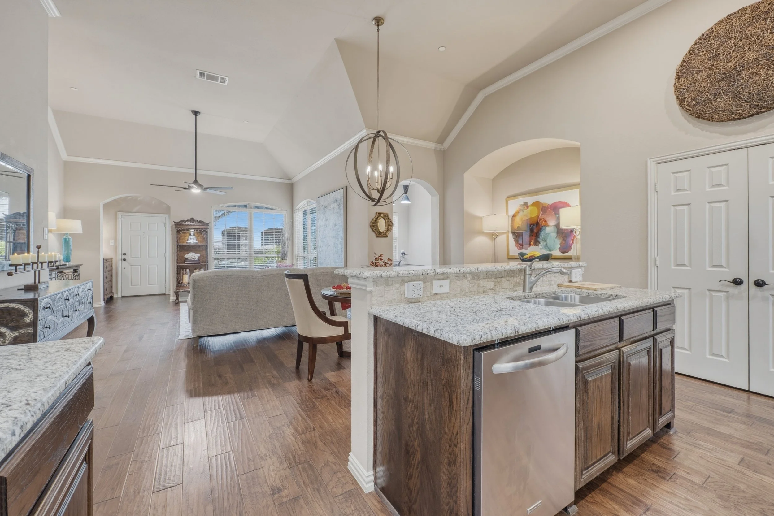 Open-concept living room and kitchen with large windows, hardwood floors, beige walls, and modern furniture including a couch, dining table, and decorative art, with light fixtures hanging from the ceiling.