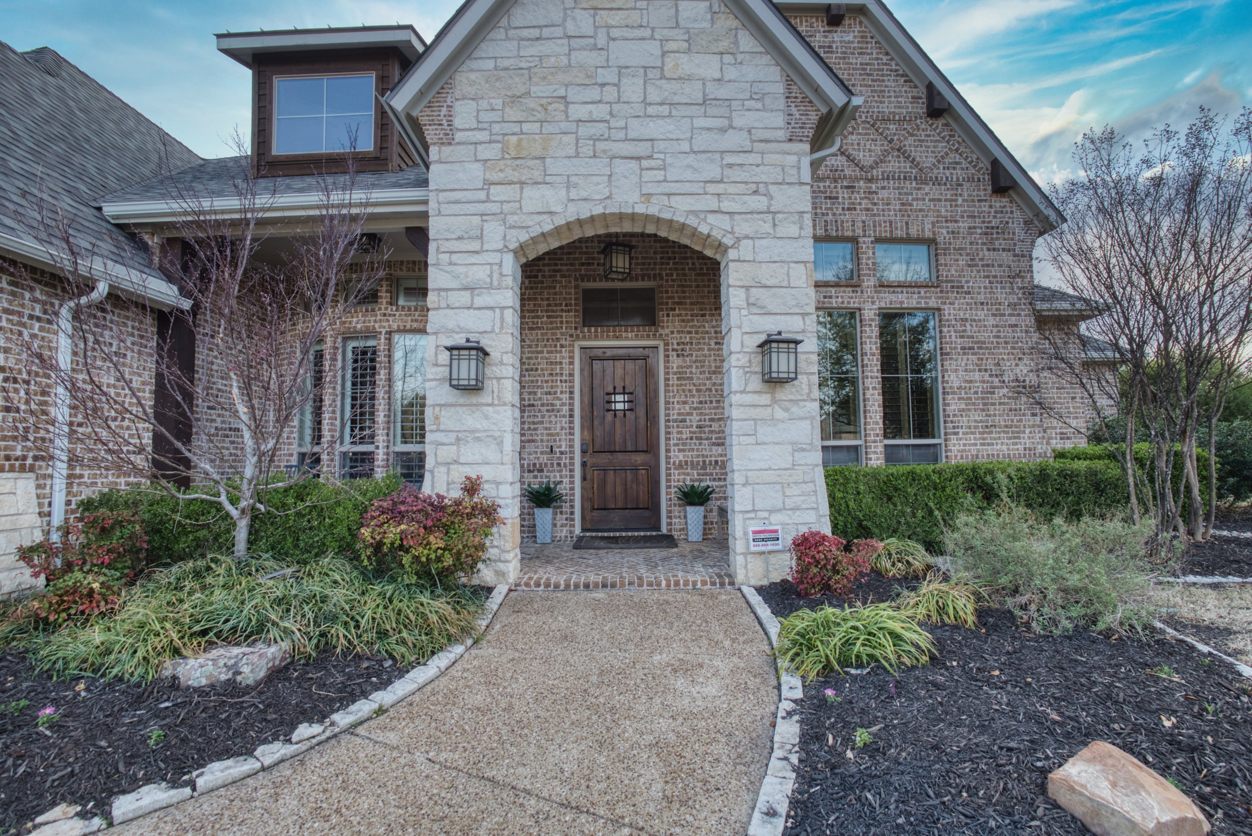 Front view of a house with a brick and stone facade, wooden front door, and landscaped yard with plants and trees.