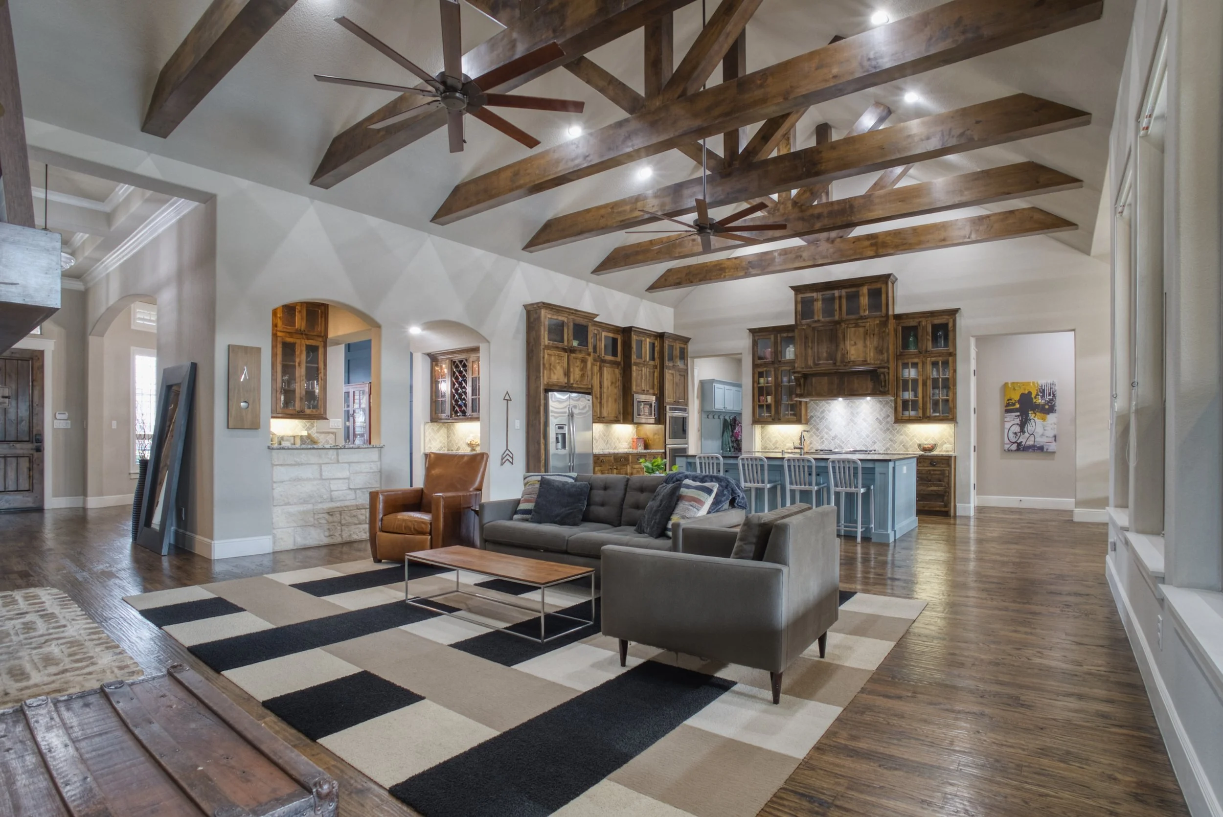 Spacious open-concept living room with high vaulted ceiling featuring exposed wooden beams and ceiling fans. A gray sofa with cushions, a tan armchair, and a coffee table sit on a geometric patterned rug. The background displays a kitchen with wooden