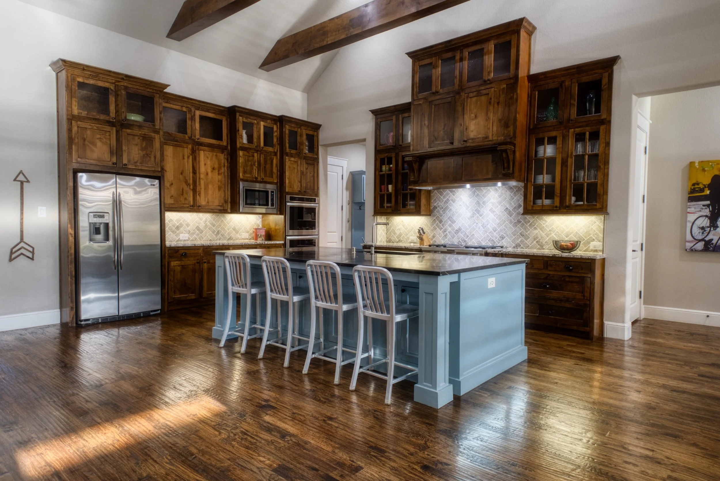 Modern kitchen with wooden cabinets, stainless steel refrigerator, and a light blue island with five bar stools, illuminated backsplash, and wood-beamed ceiling.