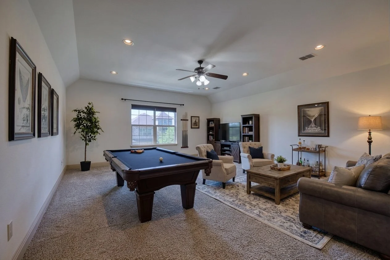 Living room with a pool table, beige carpet, and white walls. Features include a ceiling fan, large window, seating area with sofas and armchairs, a wooden coffee table, TV, and entertainment center. Decor includes framed artwork and a tall potted pl