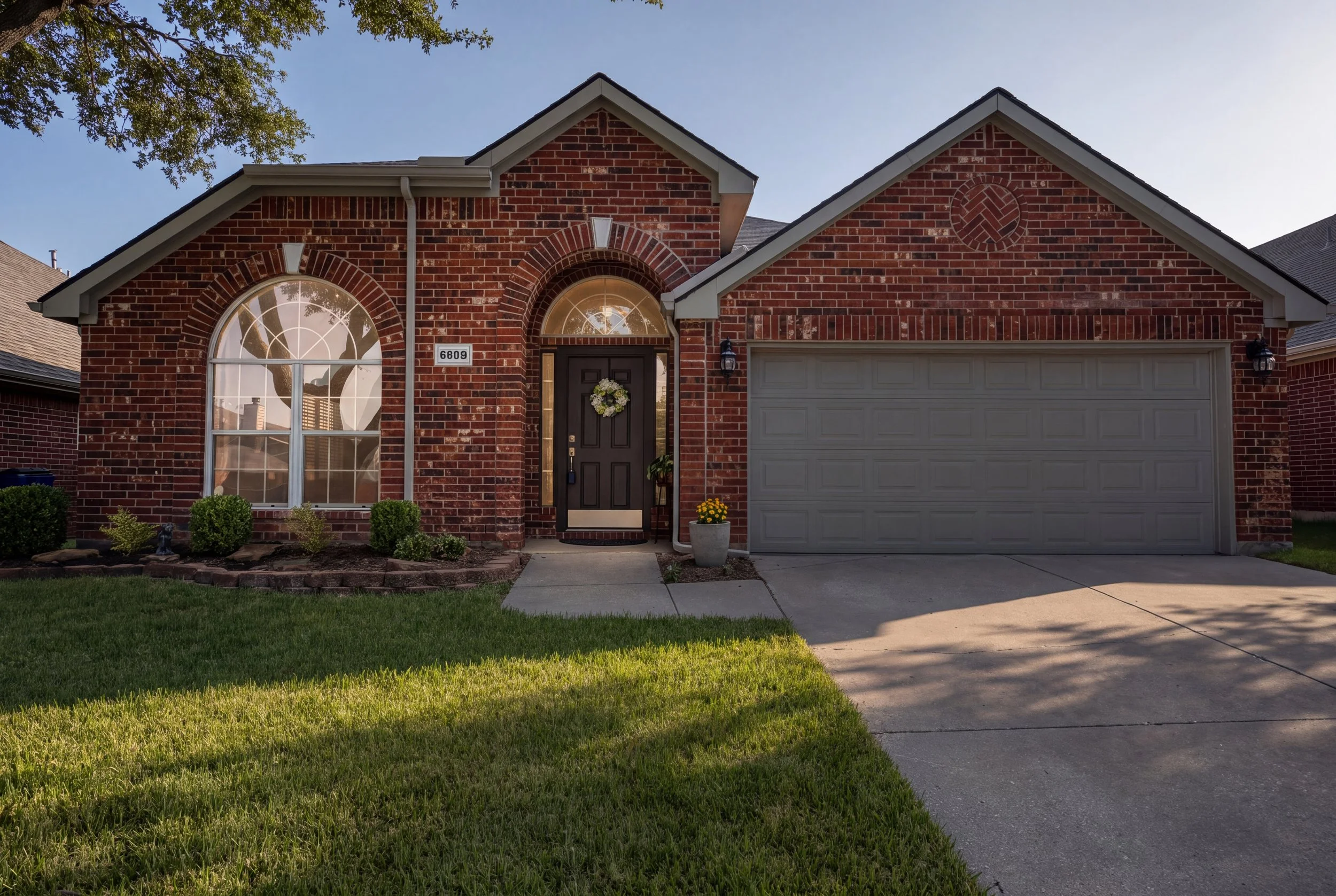 A brick house with an arched window, front door decorated with a wreath, and a small garden in a residential neighborhood.