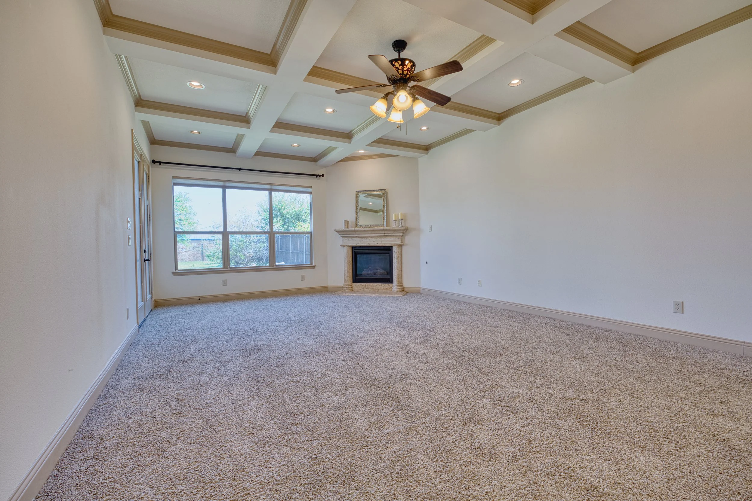 Empty living room with beige carpet, white walls, large window, ceiling fan, and fireplace with mirror above it.