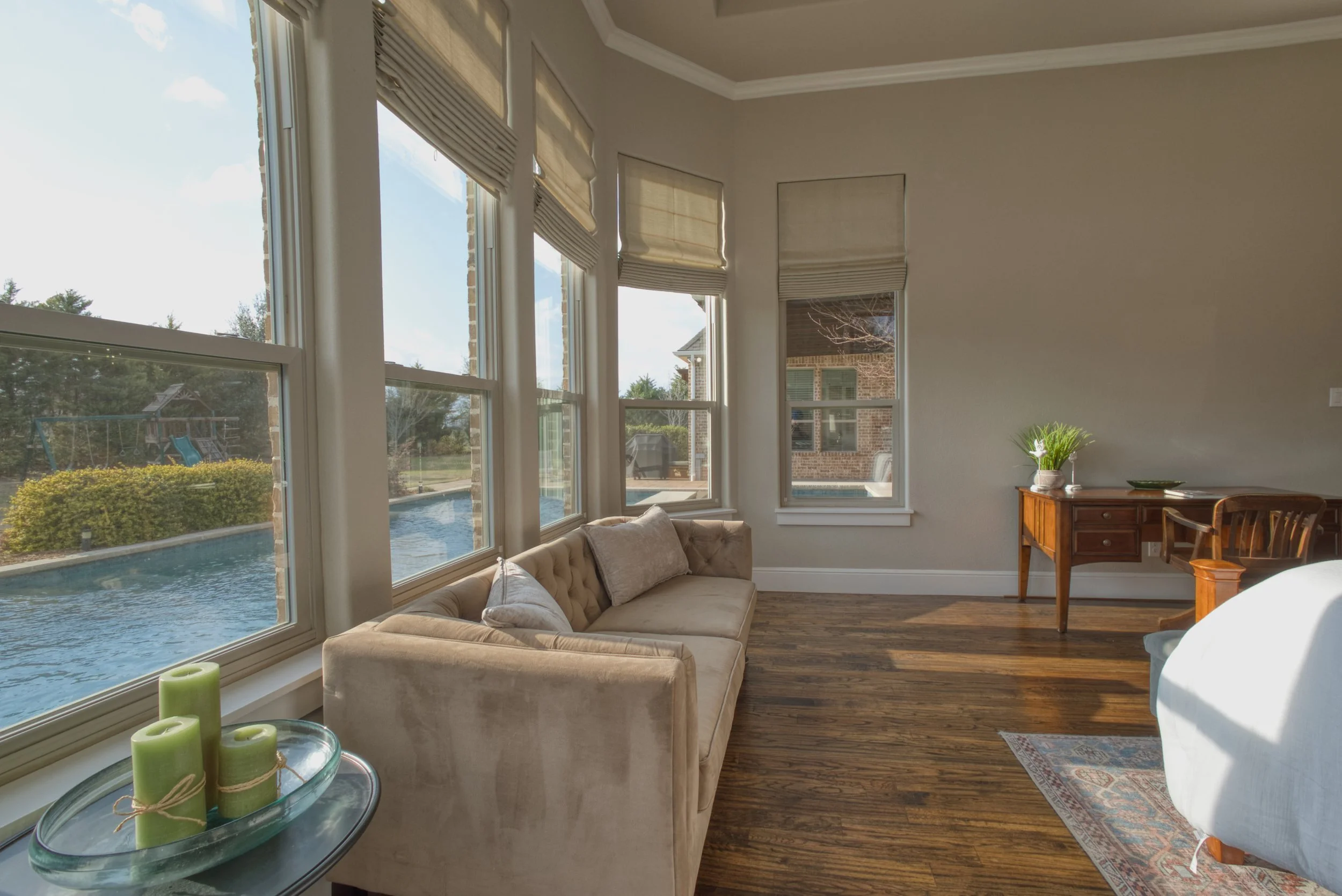 Living room with large windows overlooking a backyard with a swimming pool, a swing set, and bushes. Contains a beige tufted sofa with pillows, a glass table with green candles, a small wooden desk with plants, and a white armchair.