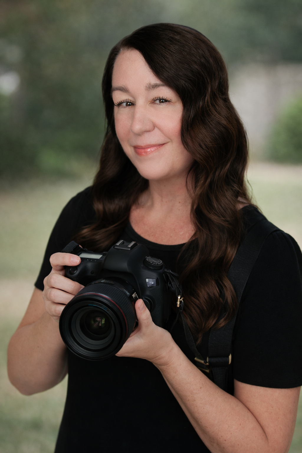 A woman with long wavy brown hair smiling and holding a professional camera outdoors.