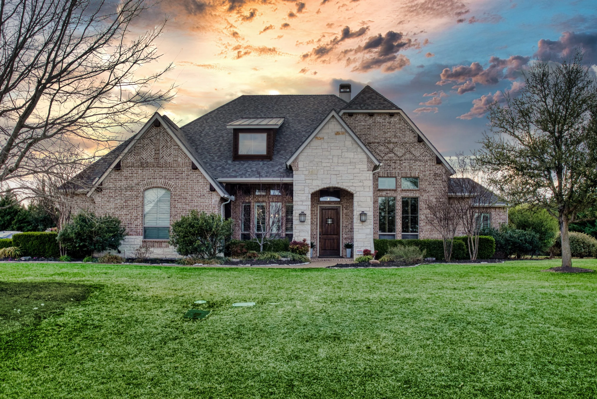 Front view of a two-story brick house with a landscaped yard and trees at sunset, featuring a wooden front door and multiple windows.