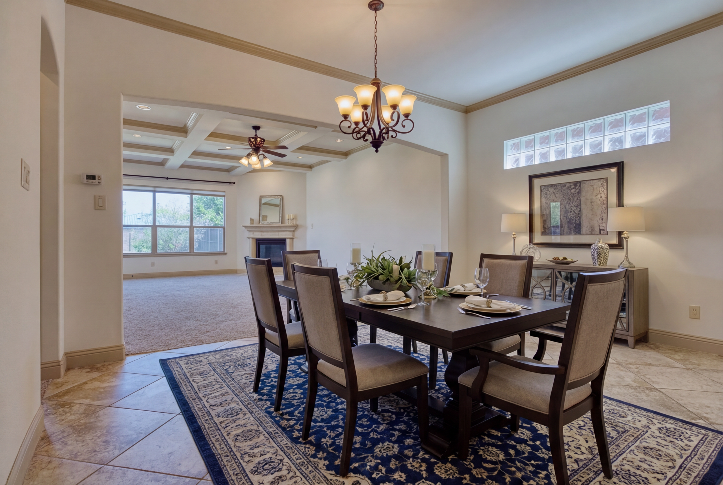 Dining room with a dark wood table set with plates, glasses, and candles, surrounded by eight beige upholstered chairs. A chandelier hangs above. Adjacent living space with a large window, a ceiling fan, and a fireplace. Decor includes a framed pictu