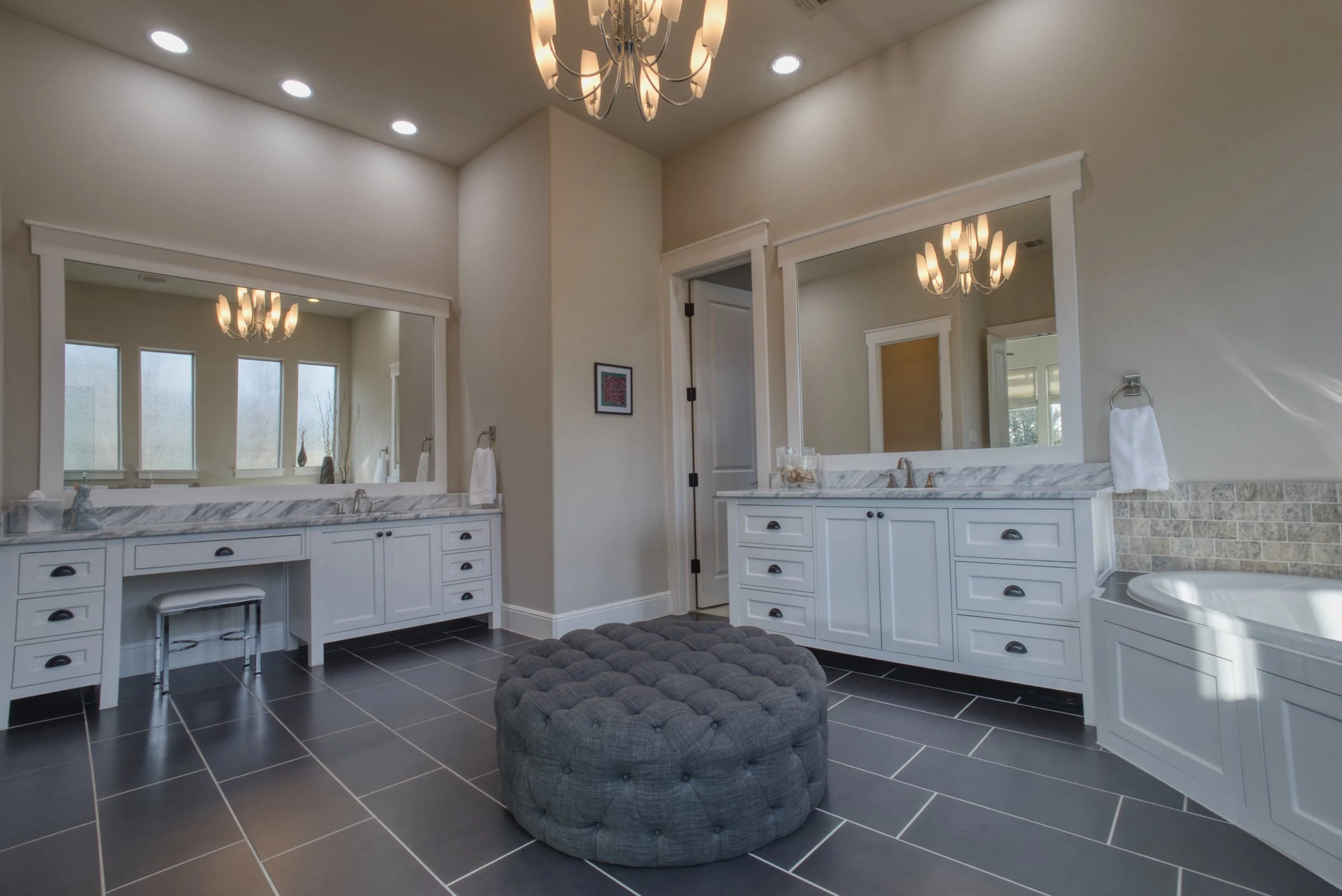 A spacious bathroom with two large mirrors above white vanities, black hardware, marble countertops, and a tiled black floor. There is a tufted round gray ottoman in the center, a chandelier hanging from the ceiling, and three small windows allowing 