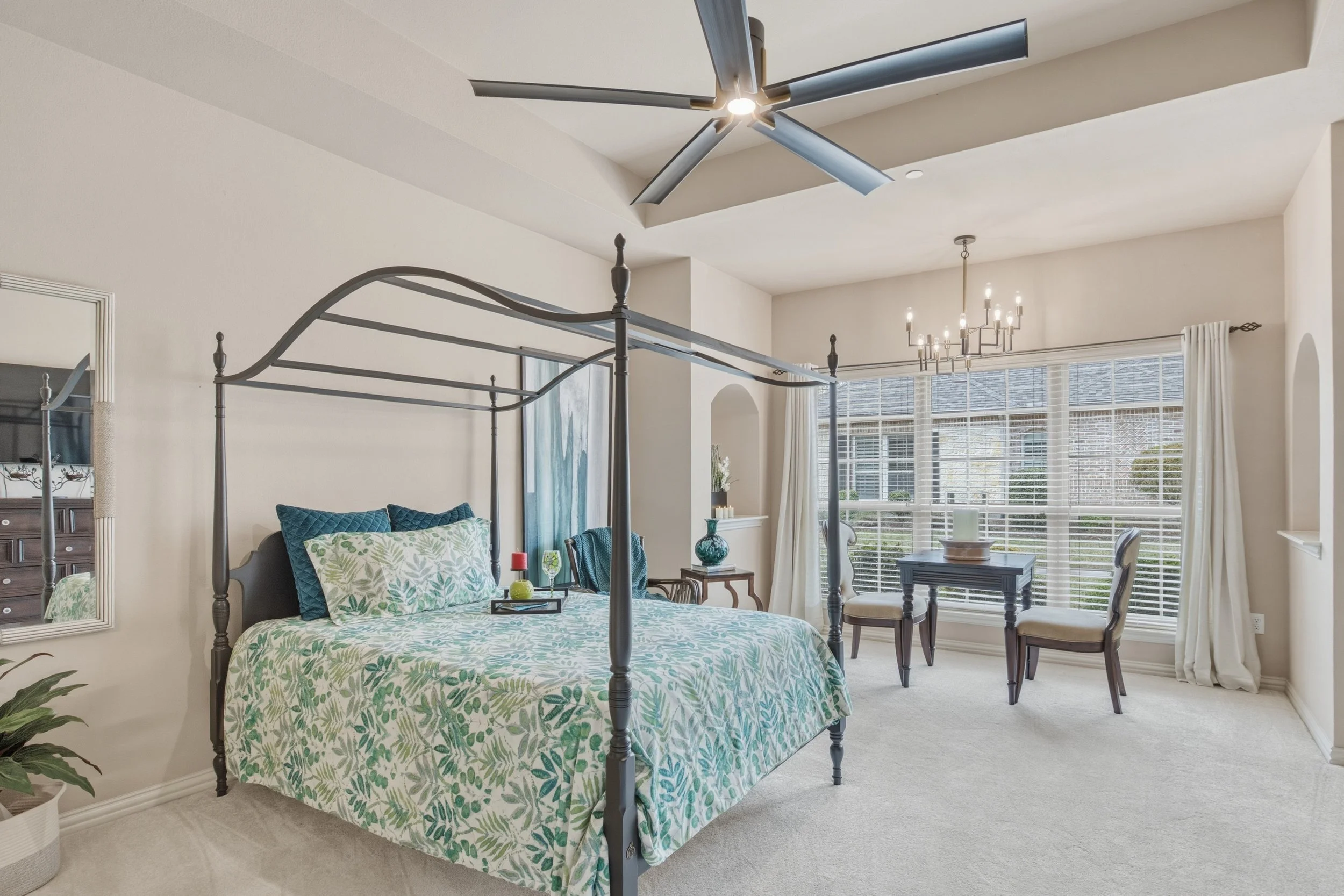 Bright bedroom with a four-poster bed, patterned bedding, and pillows, a mirror on the left, a ceiling fan above, and a sitting area with a small table and chairs by large windows with white curtains.