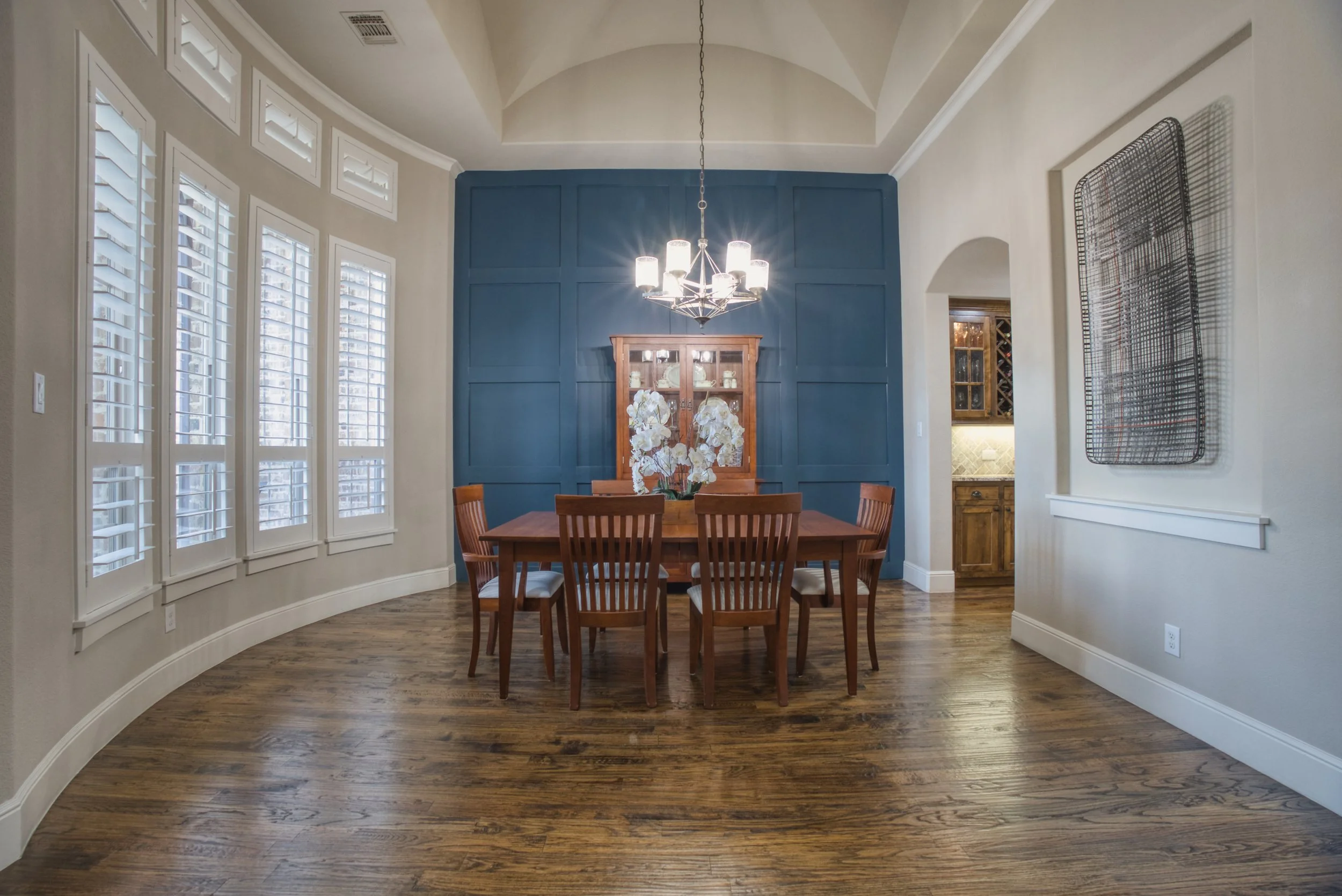 Dining room with wooden table and chairs, blue accent wall, chandelier, floral centerpiece, large window with shutters, and artwork on the wall.