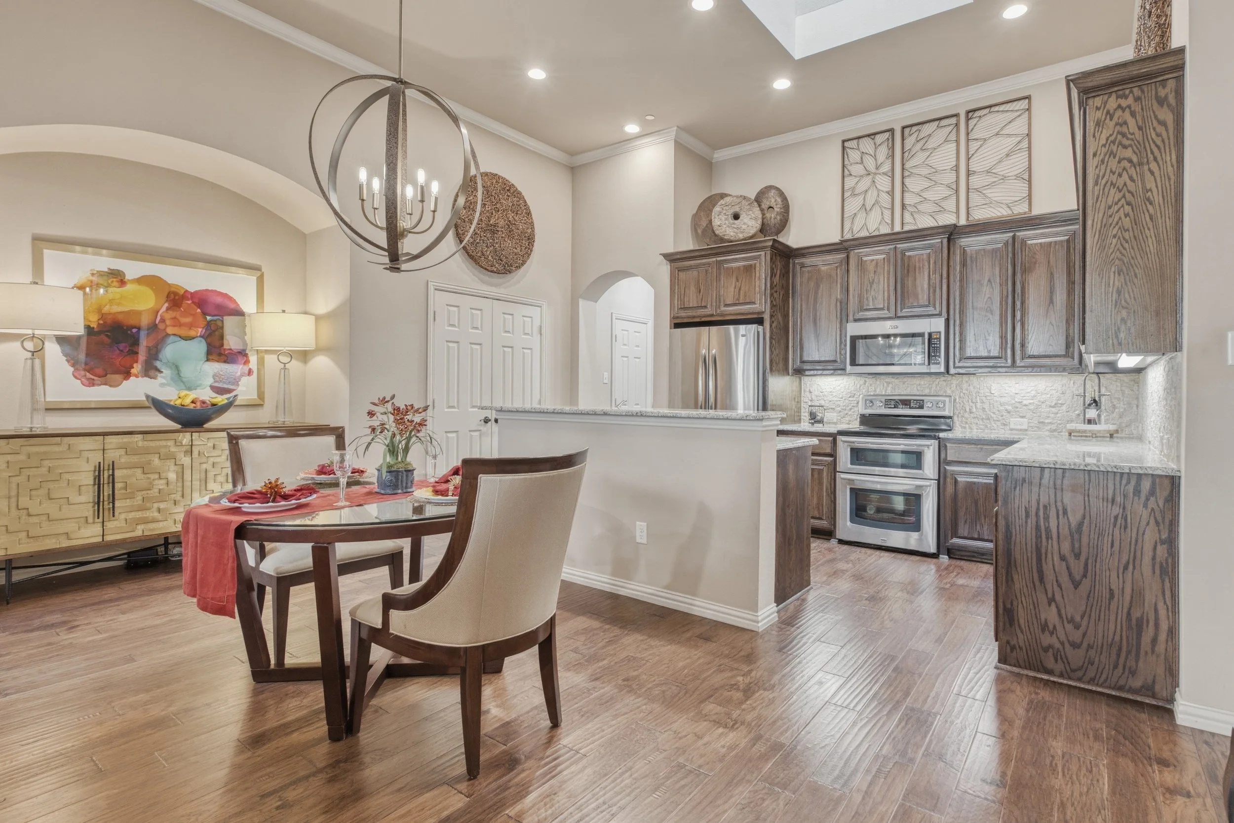 Open concept kitchen and dining area with wooden cabinets, stainless steel appliances, a small breakfast bar, a round dining table with four chairs, a modern chandelier, and wall decor.