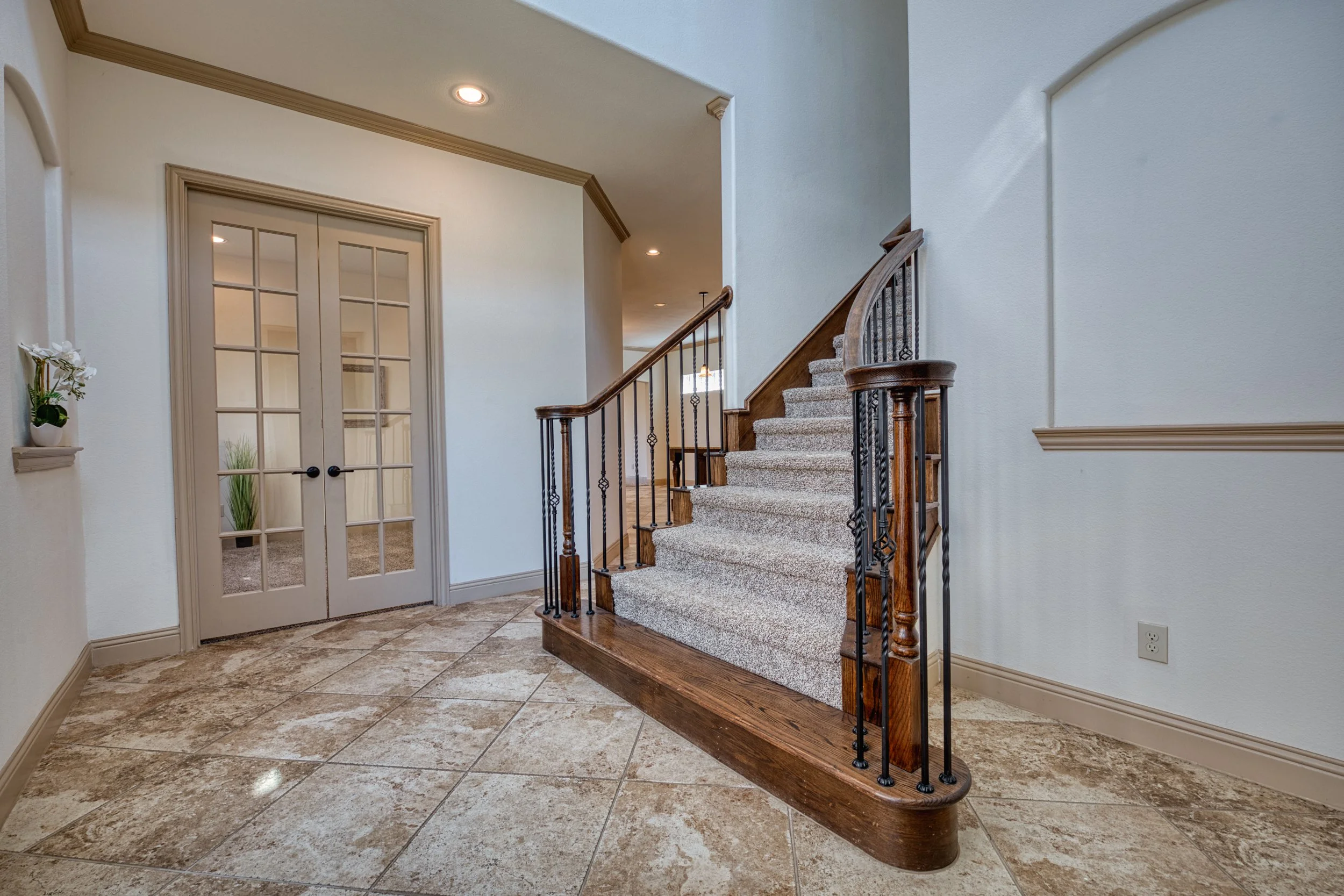 Entryway with tiled floor, staircase with carpeted steps, wooden handrail, iron balusters, glass double doors, and a small wall-mounted shelf with a potted plant.