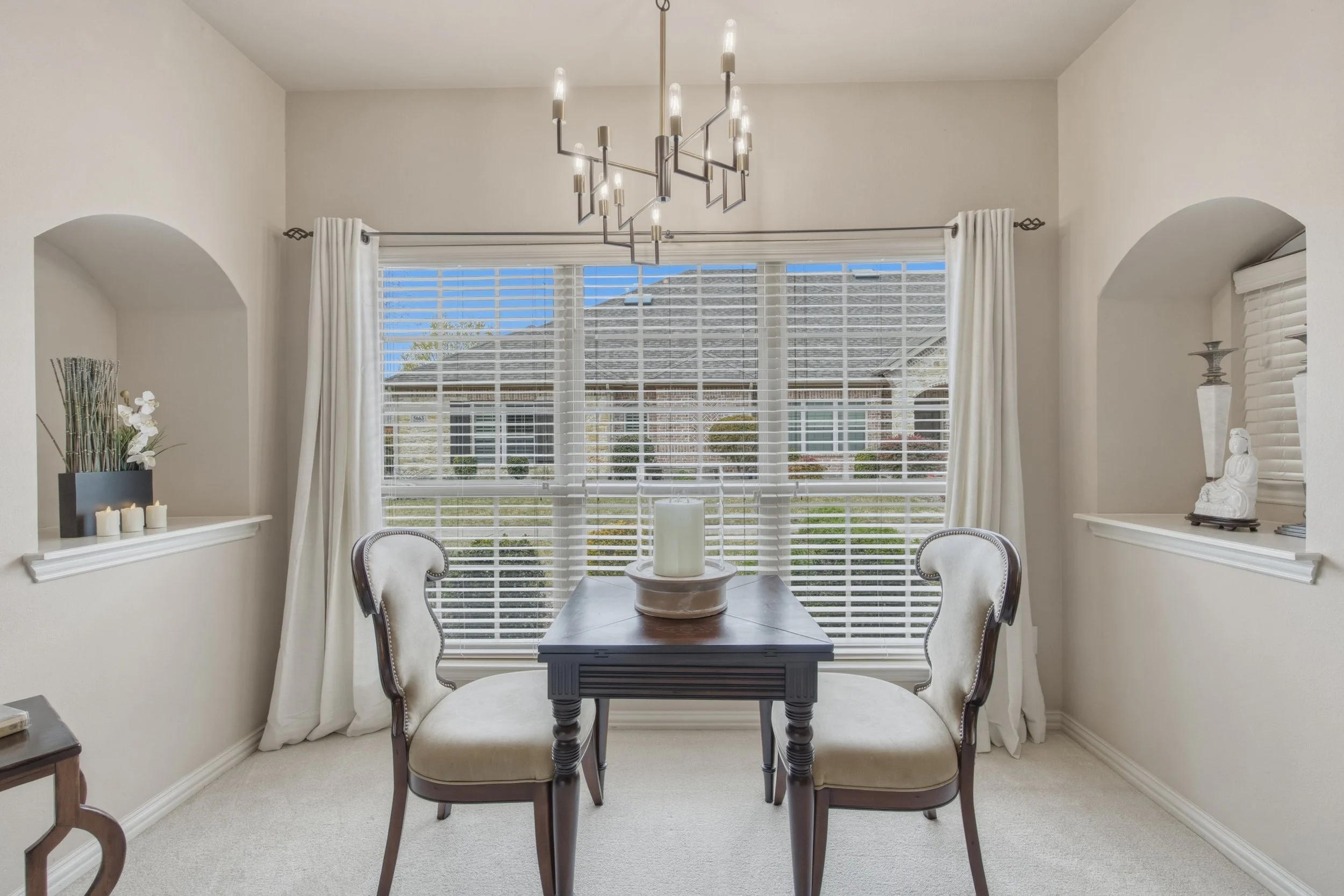 Dining room with a small wooden table, two upholstered chairs, large window with blinds and curtains, modern chandelier, decorative items on built-in shelves