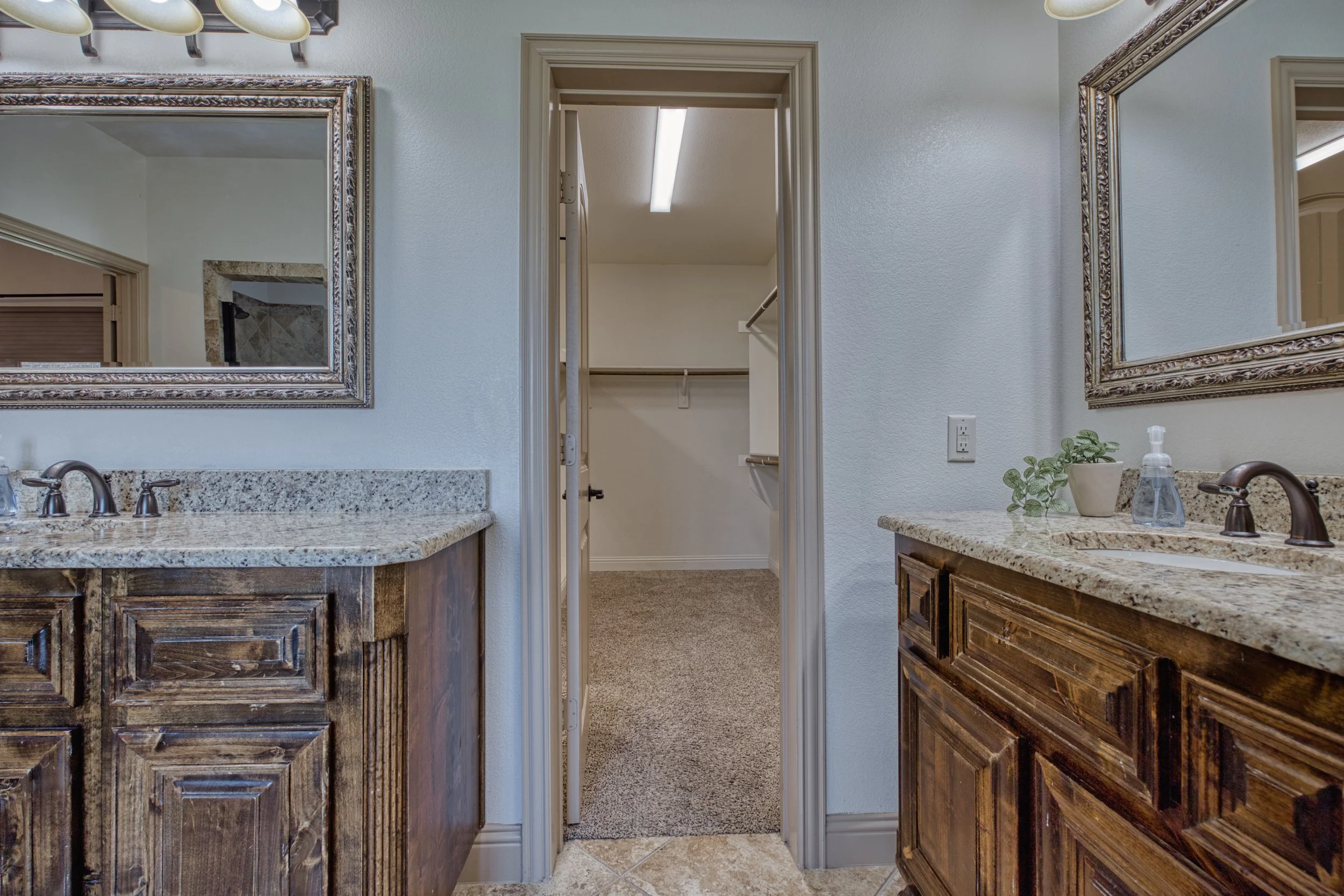 Bathroom with two wooden vanities having granite countertops and framed mirrors, a small potted plant, and a soap dispenser between the mirrors. An open doorway leads to a walk-in closet with carpeted flooring and built-in shelving.