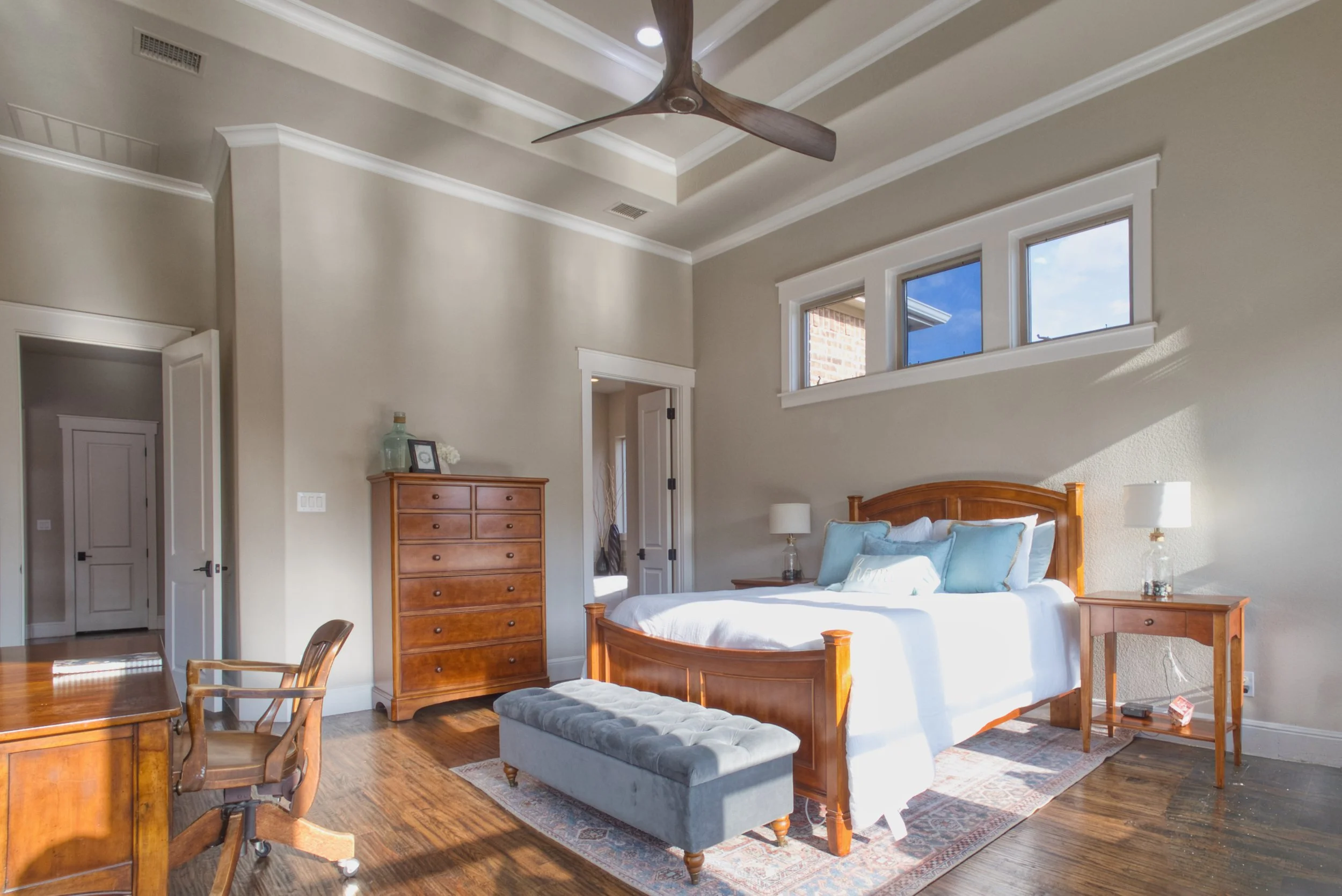 A bedroom with a wooden bed, nightstands, a tall dresser, and a gray bench. Sunlight streams through three small high windows. The room has a tray ceiling with a ceiling fan and hardwood floors.