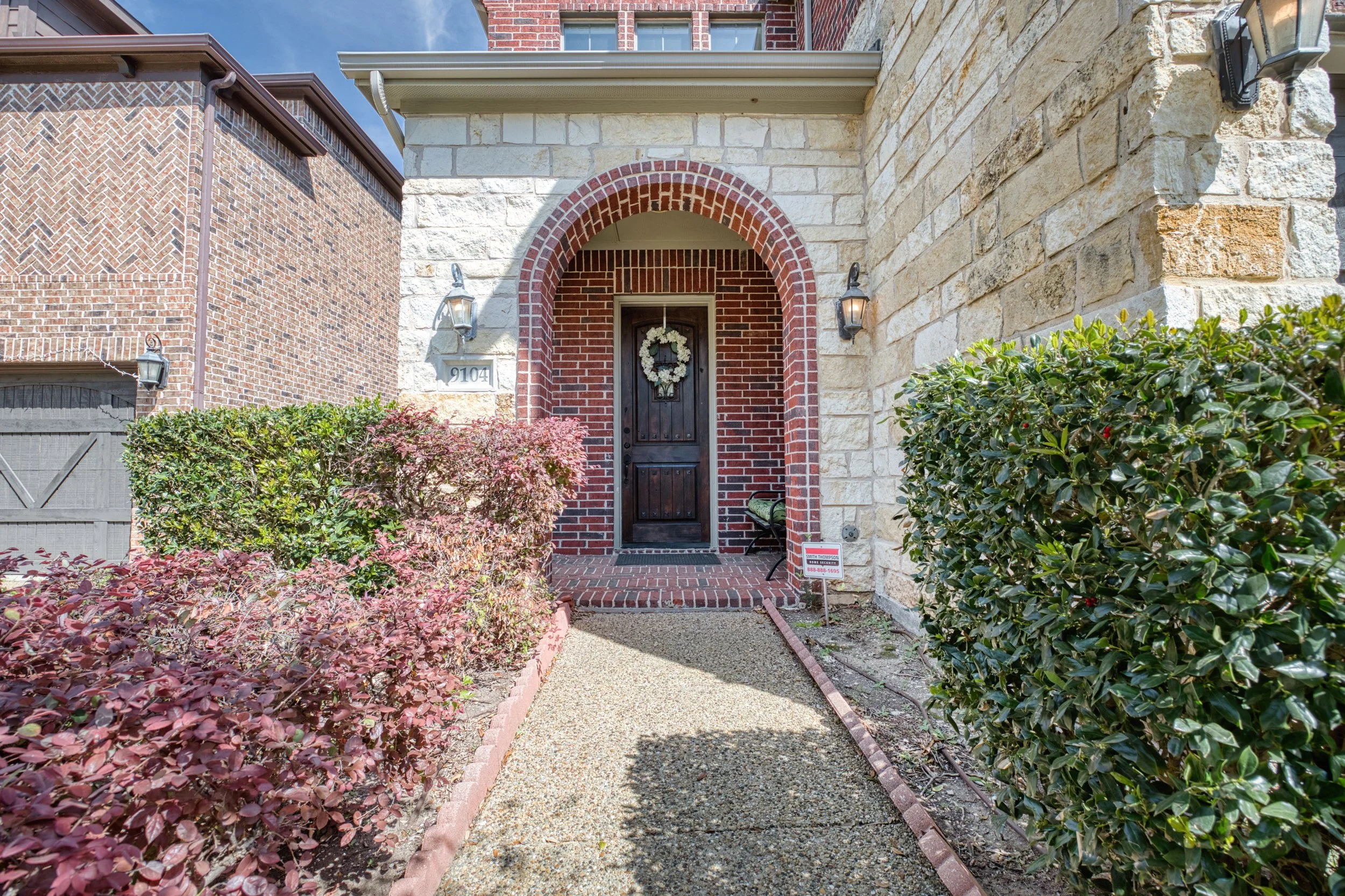 Front entrance of a house with a black door and wreath, brick and stone exterior, surrounded by green bushes and plants, with pathway leading to door.