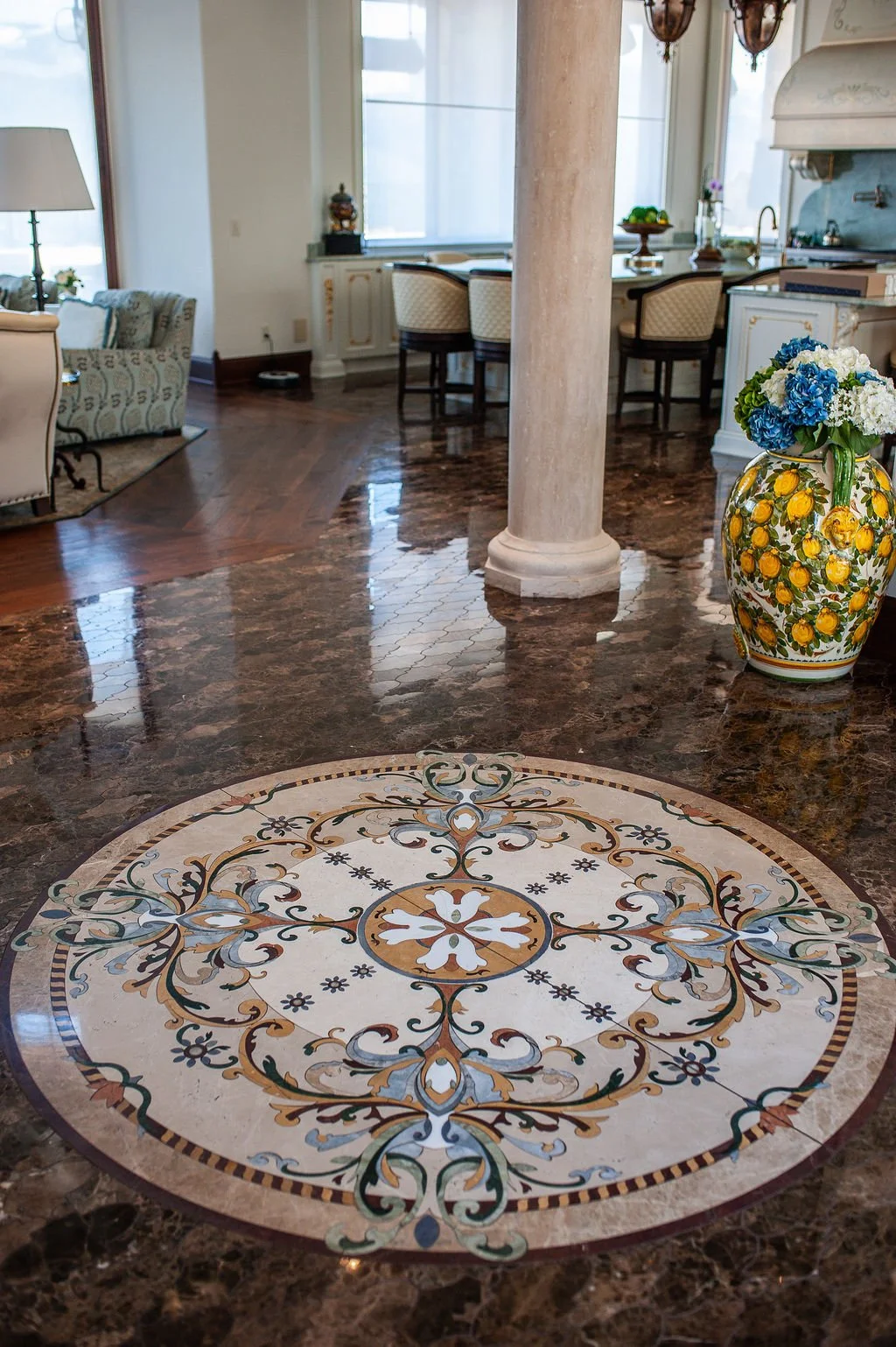 Elegant living room with a decorative mosaic tile on a brown marble floor, a large ceramic vase with blue and white flowers, a beige column, and in the background, a kitchen area with a white counter and chairs.