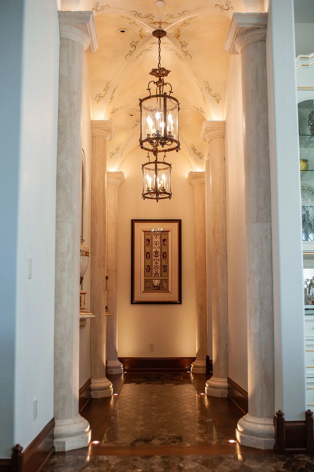 Elegant hallway with tall marble columns, a vaulted ceiling with ornate gold designs, a hanging chandelier with three lantern-style lights, framed artwork on the wall, and wooden flooring with small lights illuminating the base of the columns.