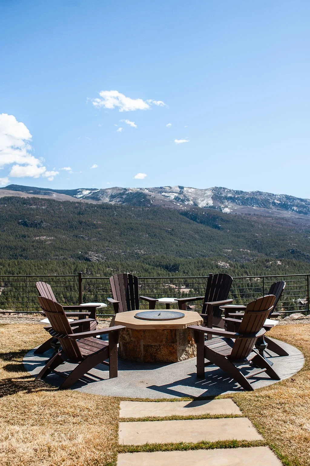 A round fire pit with six wooden Adirondack chairs around it, set on a circular concrete pad with a mountain view in the background.