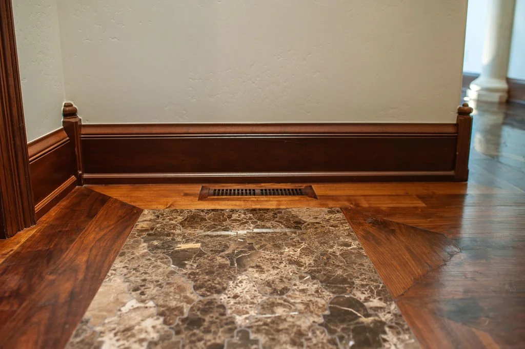 Close-up of a foyer with a marble tile floor bordered by wooden flooring, white textured wall, dark wooden baseboard, and a floor vent.
