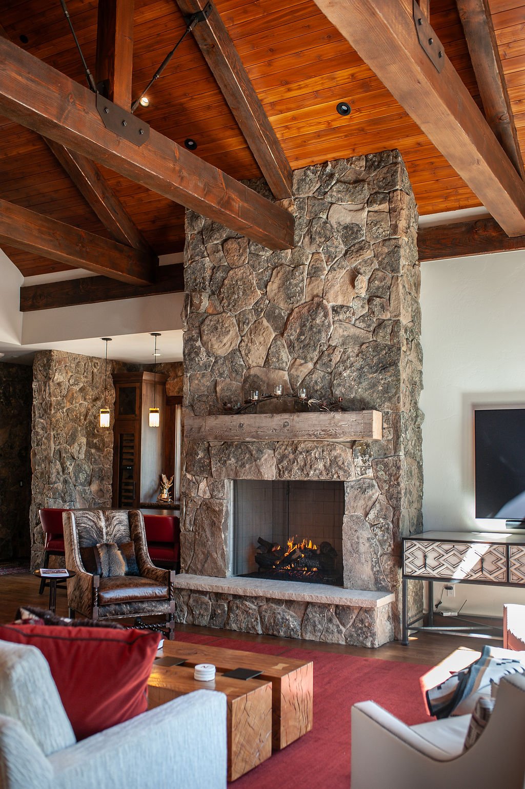Living room with a large stone fireplace, wooden ceiling beams, and modern furniture including a white sofa, a brown leather chair, and a wooden coffee table. There is a flat-screen TV on a decorative stand and a red area rug.