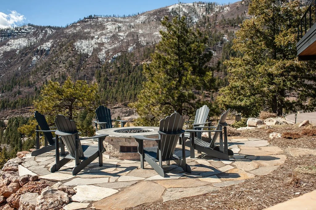 A circular outdoor fire pit surrounded by six black Adirondack chairs on a stone patio, overlooking a mountainous landscape with trees and snow-dusted peaks.