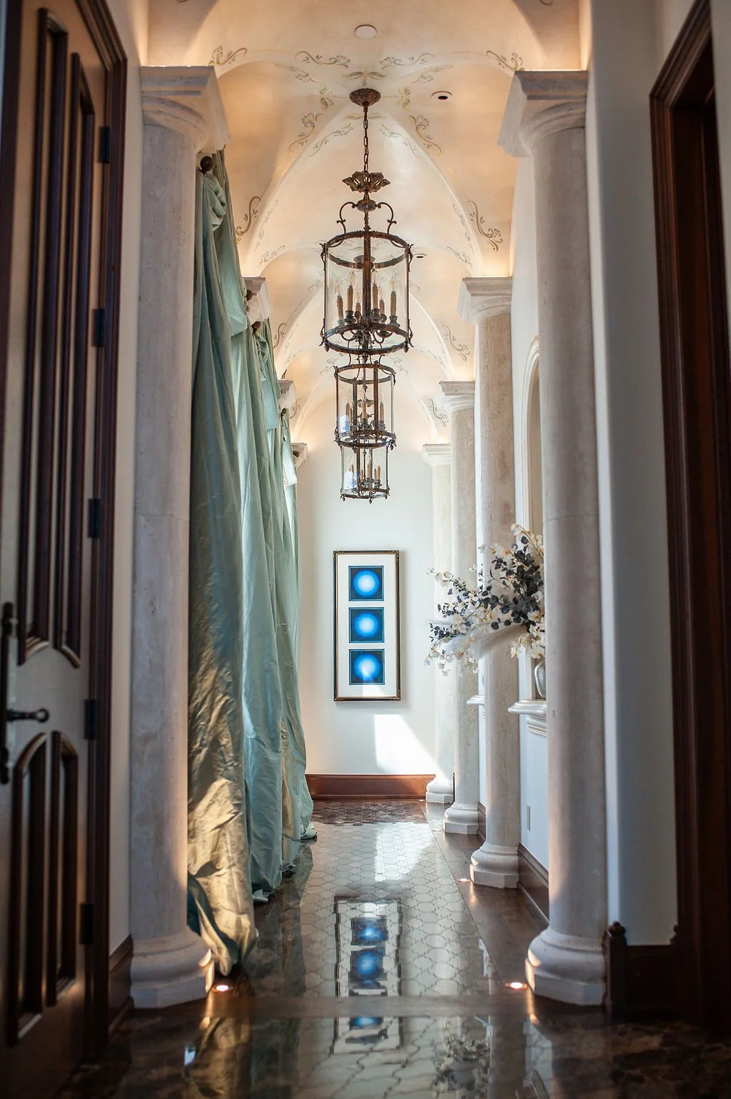 Elegant hallway with vaulted ceiling, hanging chandeliers, marble columns, framed artwork, and decorative flowers.