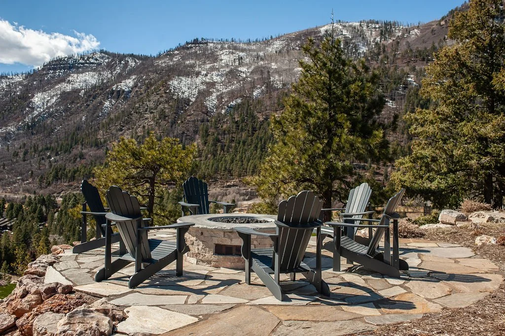 Six black Adirondack chairs arranged around a stone fire pit on a circular stone patio, with trees and mountains in the background under a partly cloudy blue sky.