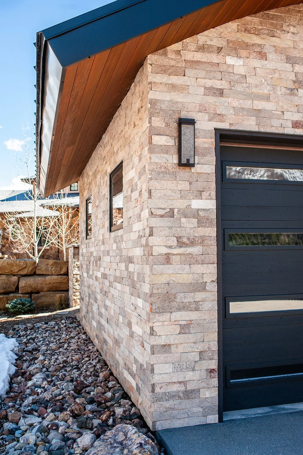 Close-up of a modern house exterior showing a stone wall with small windows, a black garage door, an exterior light fixture, and a wood-paneled roof overhang.